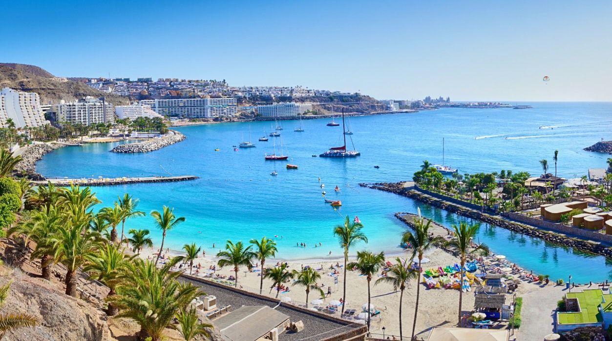 Blick auf den Strand. Blaues Wasser, Palmen, im Hintergrund große Hotels an der Promenade und kleine Schiffe im Hafen.