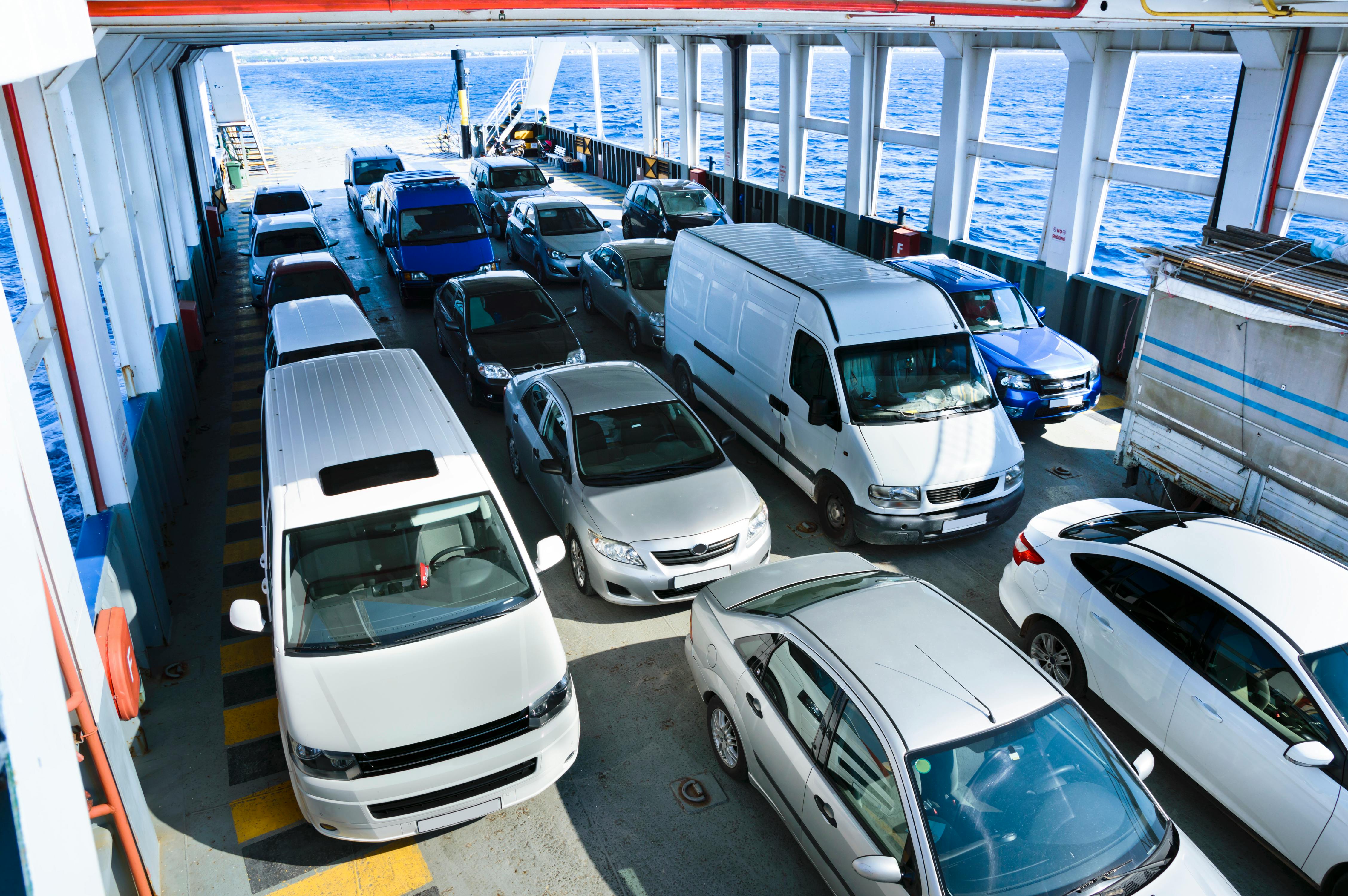 Man sieht das Fahrzeugdeck einer Fähre, auf dem Autos und Sprinter verschiedener Modelle stehen. Der Raum hat viele Fenster, durch die man das offene Meer sieht.