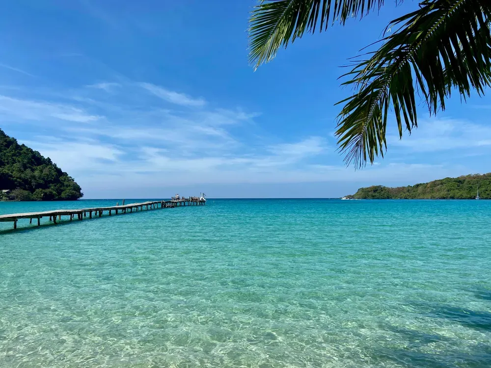 Das kristallklare Wasser am Bang Bao Beach auf Koh Kut, Thailand, glitzert in der Sonne. Palmenzweige ragen ins Bild hinein, ein scheinbar endlos langer Steg führt ins Wasser.