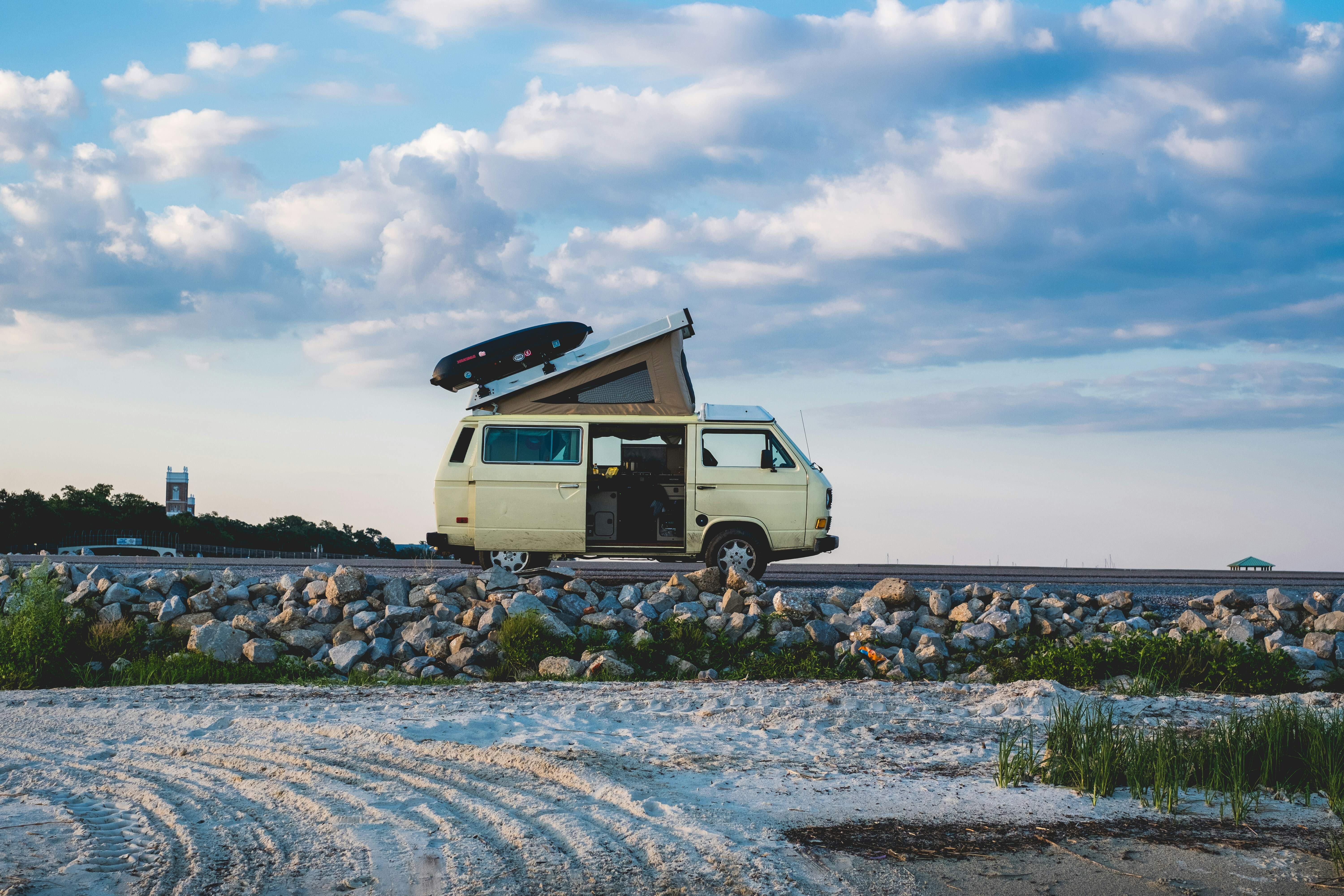 Gele camper op een weg, met op de voorgrond een zandstrand en een blauwe lucht.