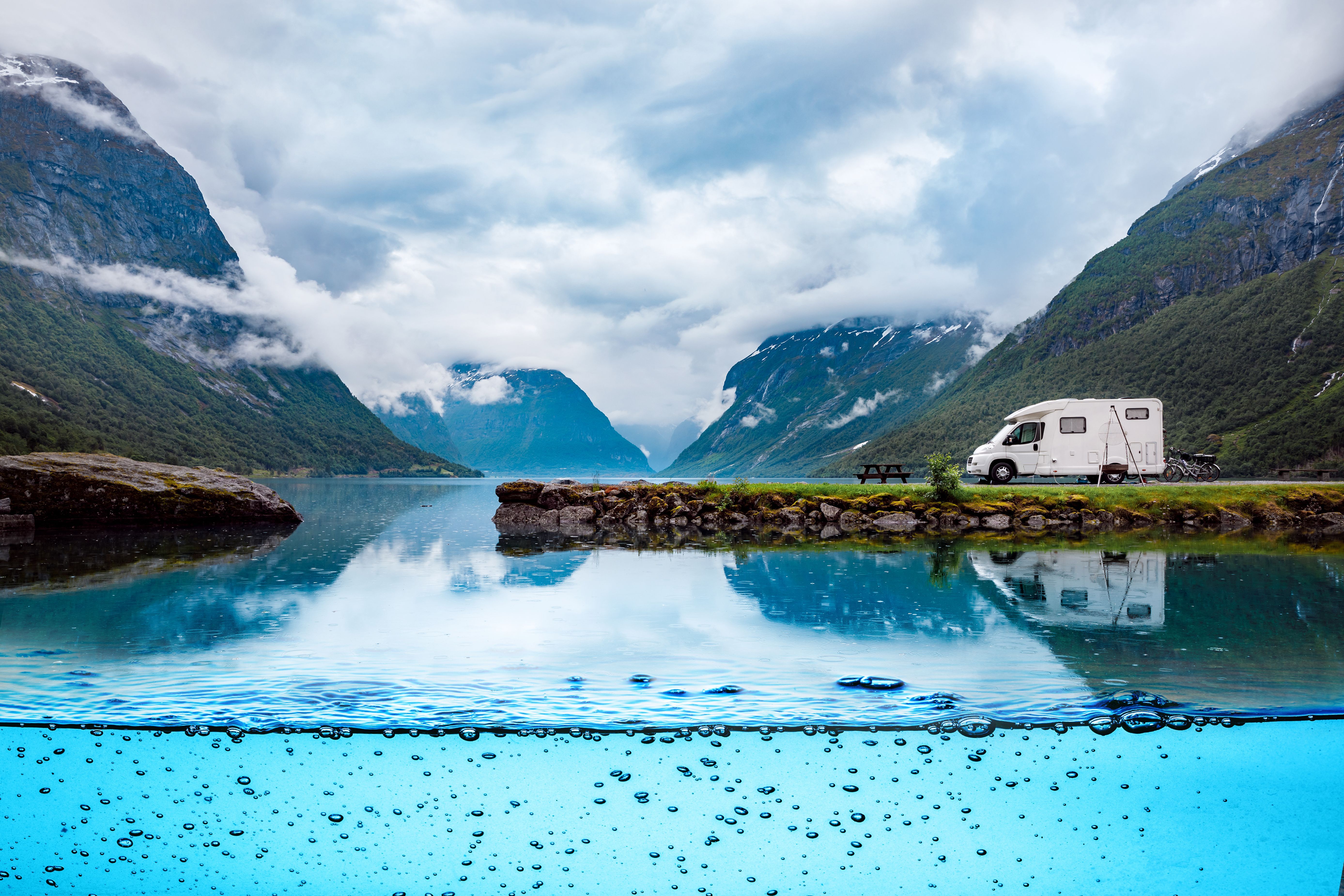 Een camper geparkeerd aan het fjord, met op de achtergrond de bergen en helderblauw water.