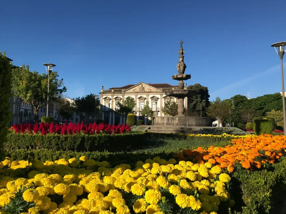Bunte Blumen in gelb, orange und rot stehen zwischen grünen Hecken hervor. Im Hintergrund sieht man ein Gebäude und einen Brunnen.