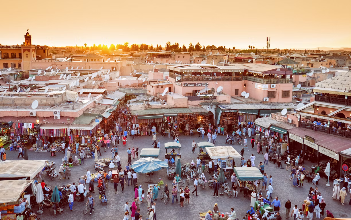 Der Platz Djemaa el-Fna in Marrakesch ist bei Sonnenuntergang belebt. Viele Menschen tummeln sich auf dem Platz mit ein paar Ständen, im Hintergrund sieht man die Dächer von Marrakesch.