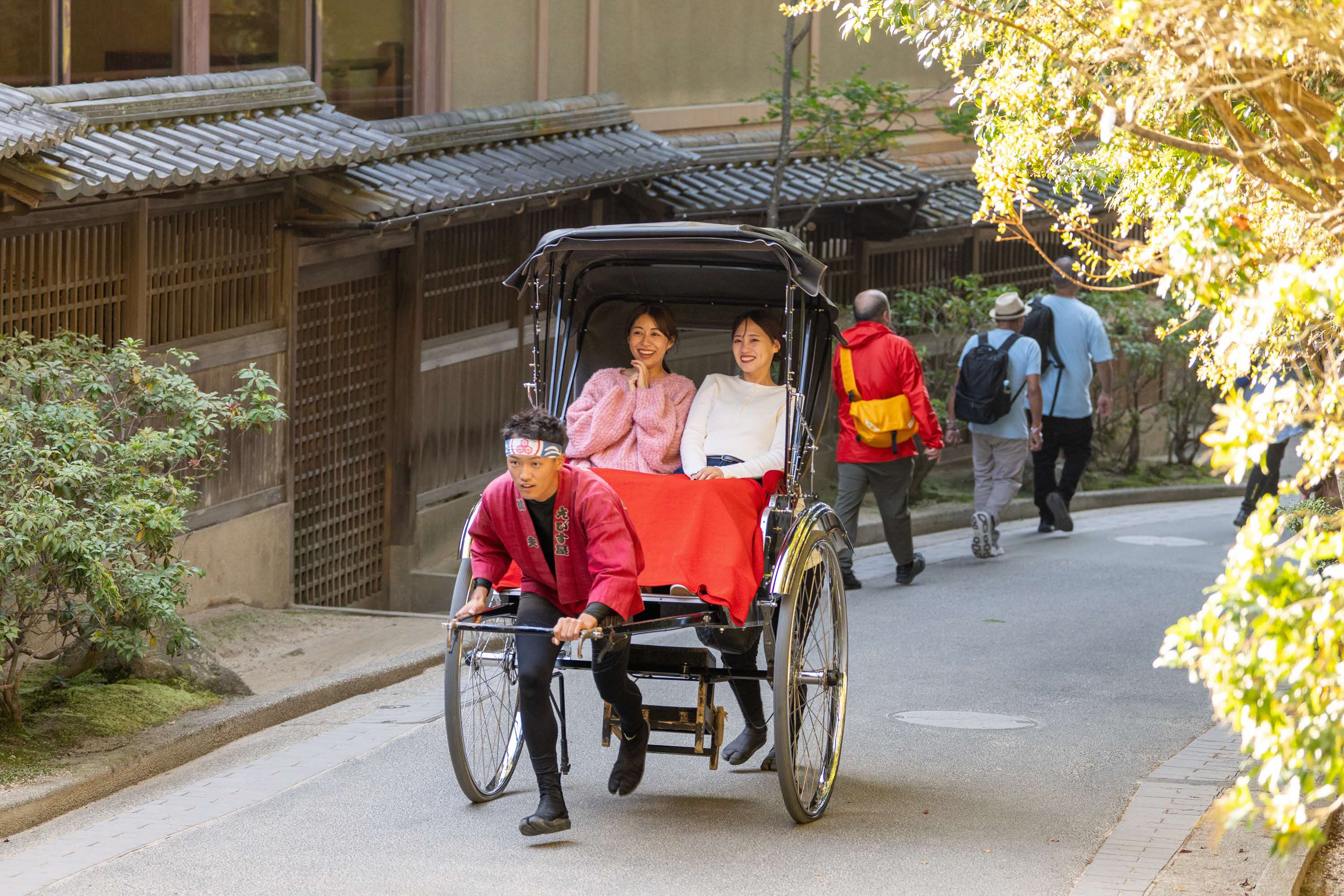 A person dragging a traditional cart with two women sitting in it up a traditional Japanese street.