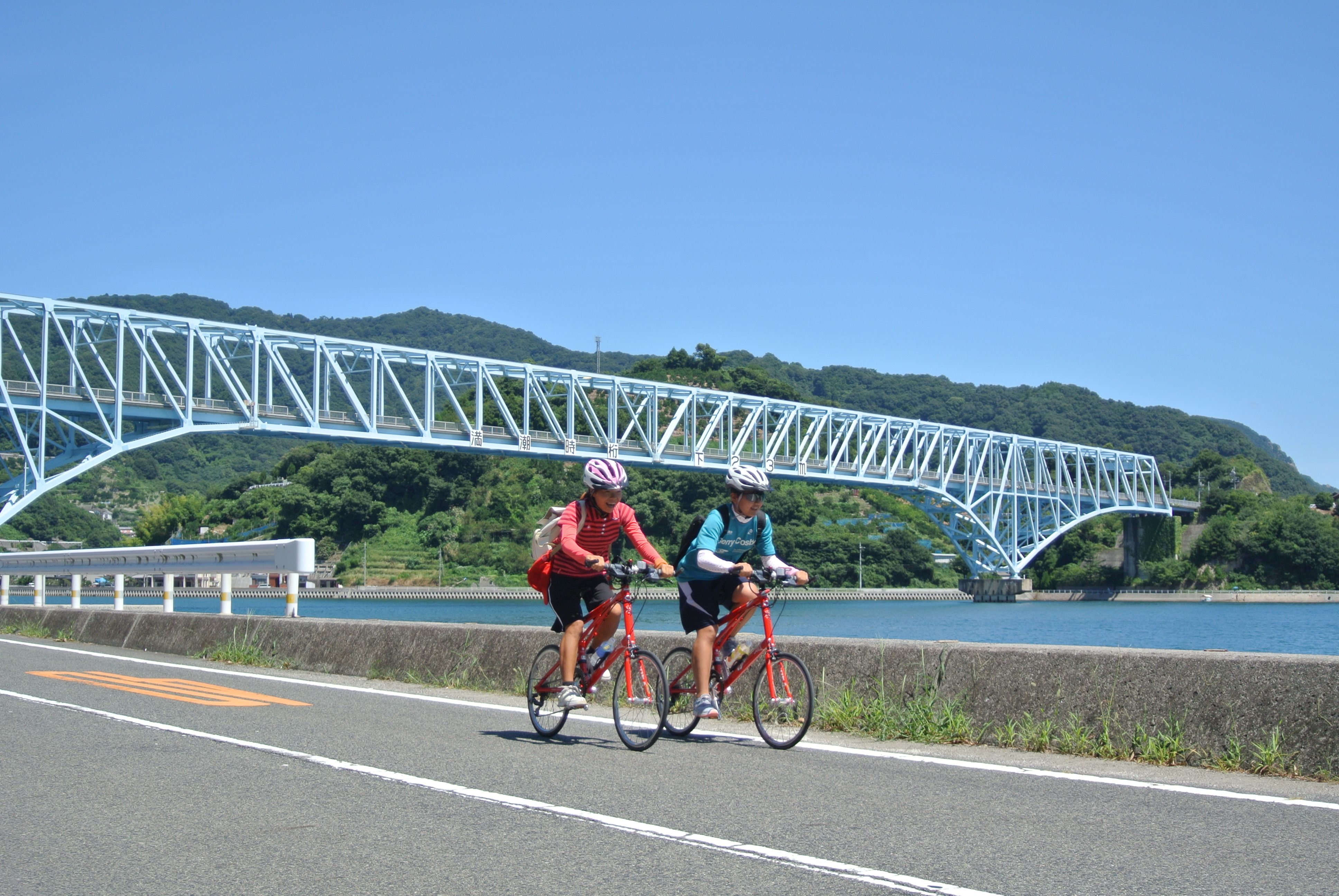 Two people cycling down a road, a bridge and river in the background.