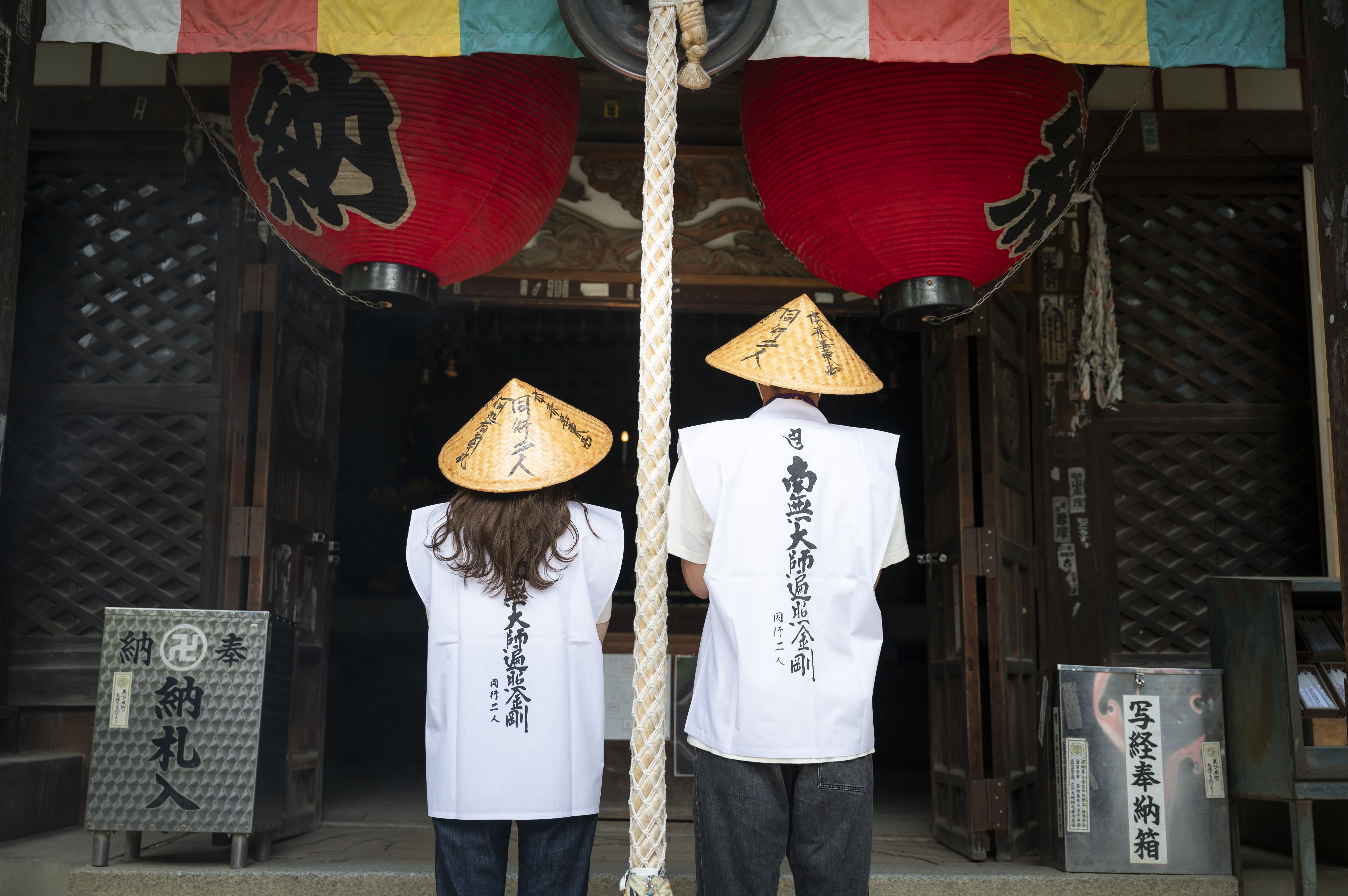 Deux personnes vues de dos, vêtues de costumes traditionnels et coiffées de chapeaux, dans un temple.