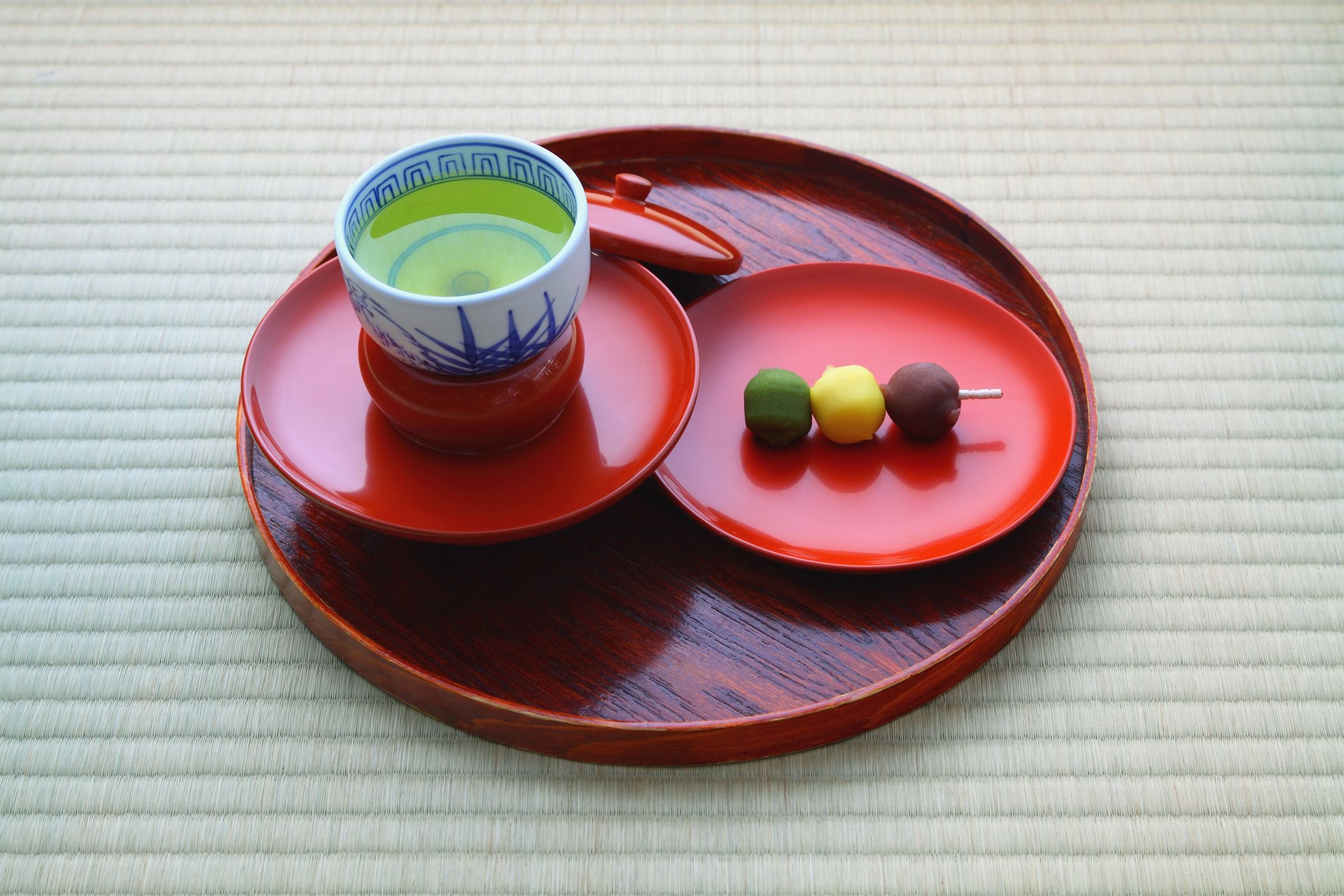 Auf einem rötlichen Tablett stehen zwei kleine Teller. Auf einem ist grüner Tee in einer traditionell japanischen Tasse, auf dem anderen sind typisch japanische Süßigkeiten, Botchan dango.
