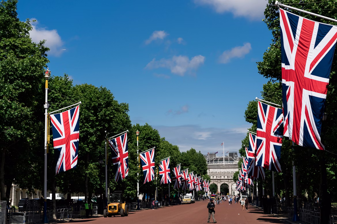 Entlang der Prachtstraße The Mall am Buckingham Palace wehen Flaggen des Union Jack im Wind. Die Sonne scheint.