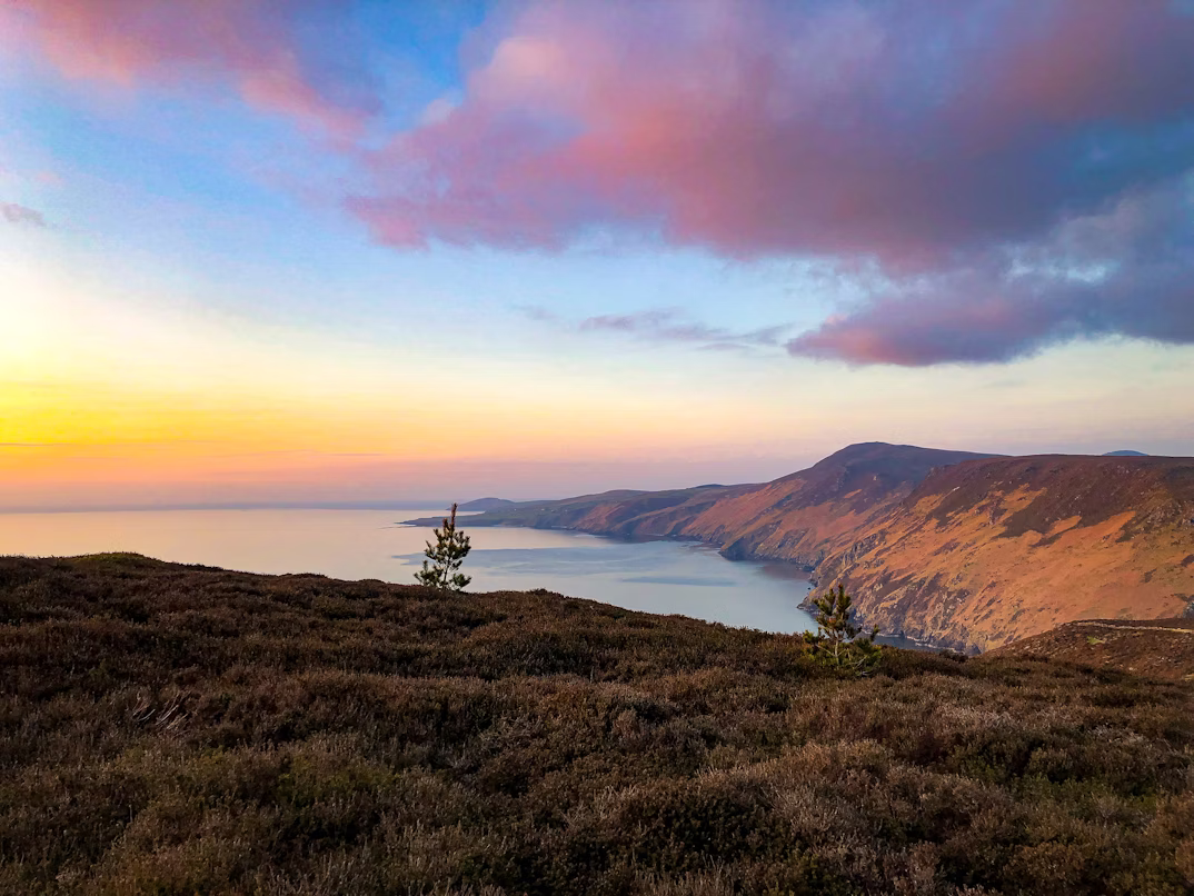 Blick von einem Hügel aus über die Bucht Fleshwick Bay auf der Isle of Man. Die Sonne geht gerade unter, der Himmel ist in Pink- und Gelbtöne getaucht.