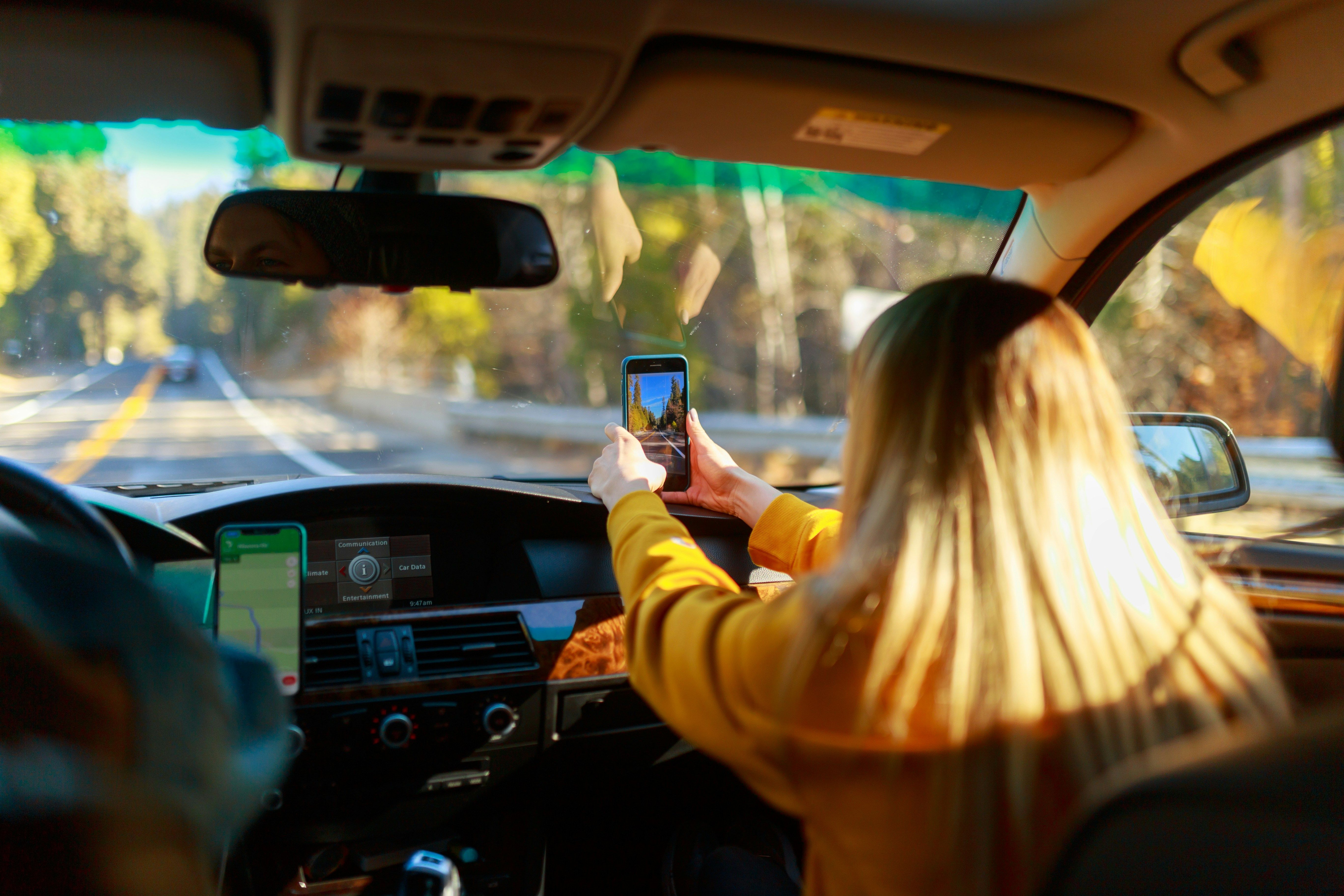 Eine Frau mit langen blonden Haaren macht mit ihrem Handy ein Foto aus dem Auto heraus.