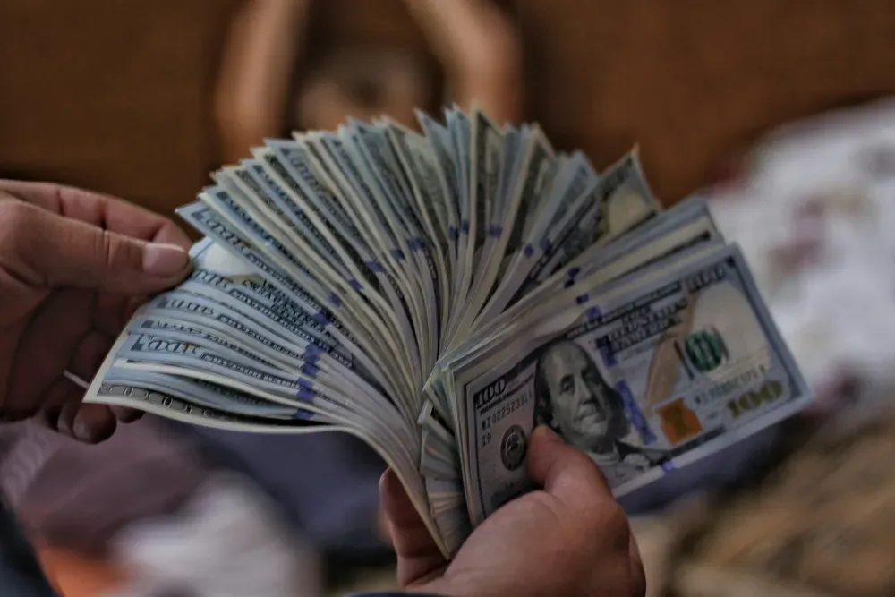 Eine Person hält zahlreiche 100-Dollar-Scheine gefächert in den Händen. Man sieht nur die Hände und die Scheine.