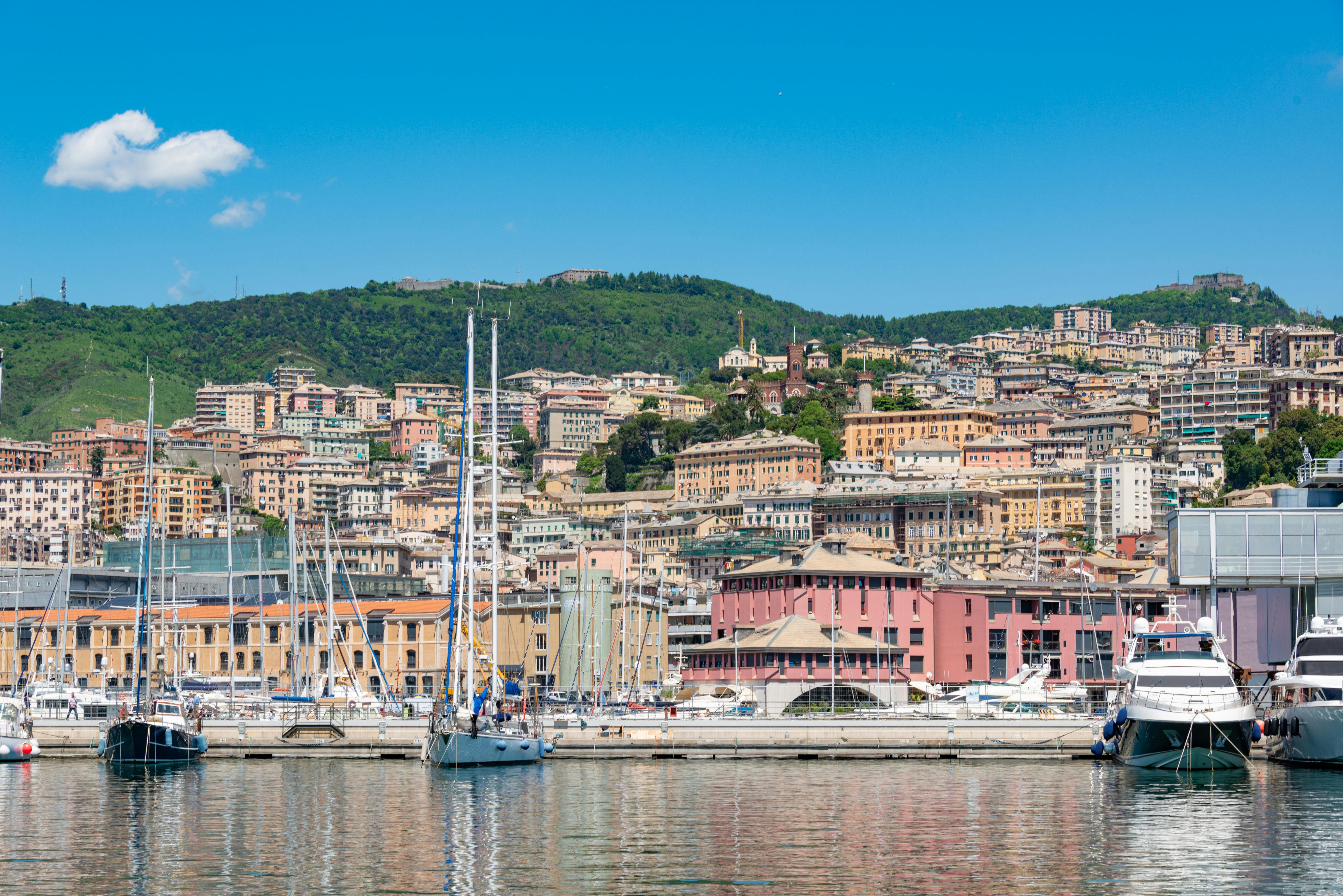 Vom Wasser aus fotografiert, sieht man einige Boote, wie sie vor der Stadtkulisse von Genua in der Sonne liegen.