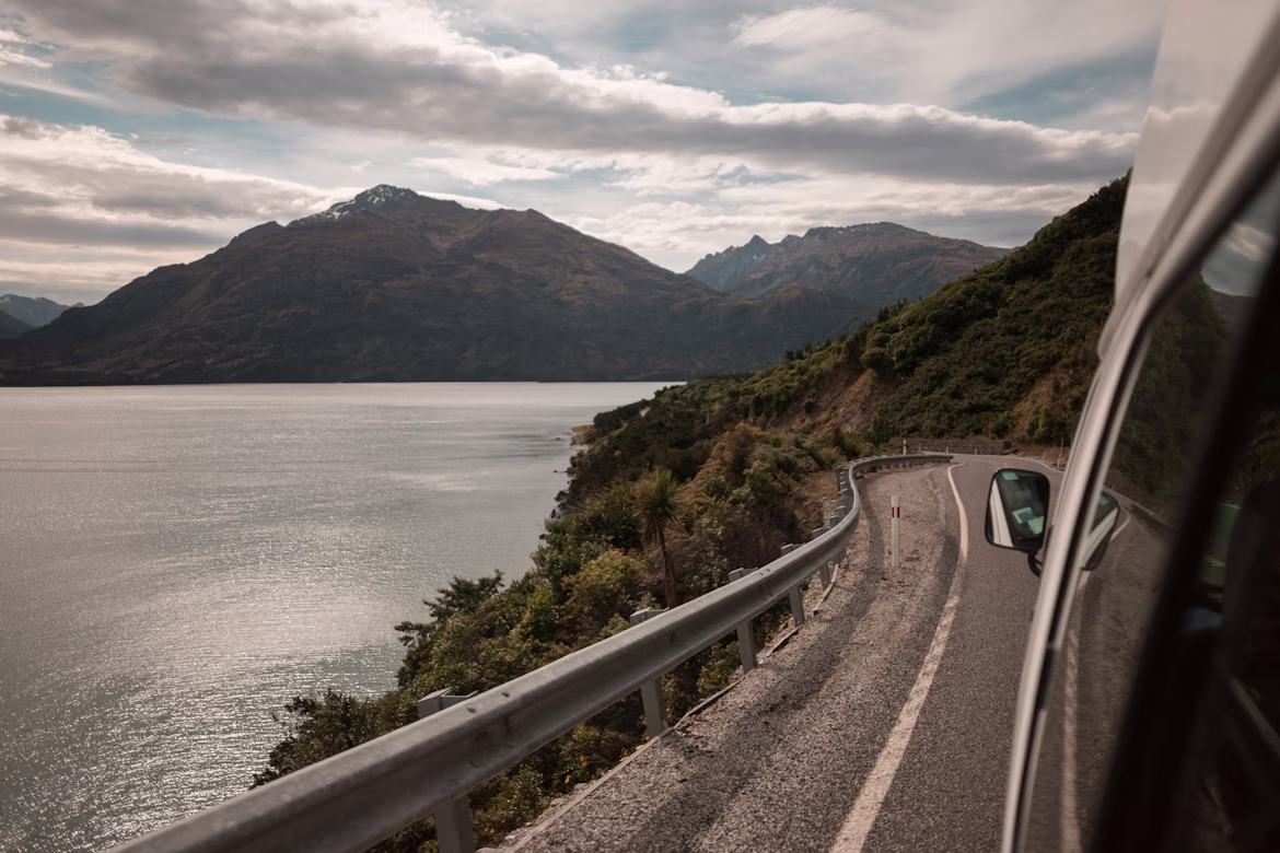 Aus einem Fahrzeug heraus fotografiert, erkennt man die malerische Fahrt entlang der Glenorchy-Queenstown Road in Neuseeland. Die Straße führt am Lake Wakatipu vorbei, im Hintergrund erkennt man die umliegende Bergkette der Remarkables.