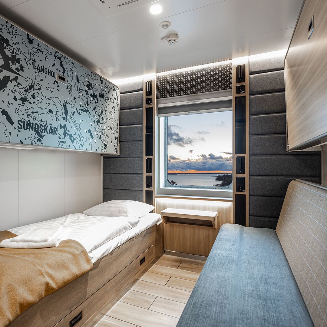 Bett links, rechts couch, an den Wänden ausklappbare Betten, hinten ein Fenster durch das das Meer zu sehen ist.