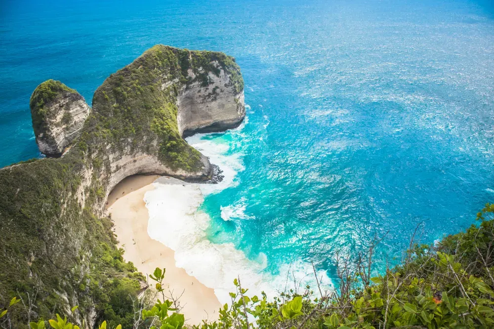 Der Kelingking Beach auf Nusa Penida sieht aus wie ein T-Rex. Die dicht bewachsenen Felsen ragen ins türkise Meer, die Wellen rauschen am Ufer des weißen Sandstrandes.