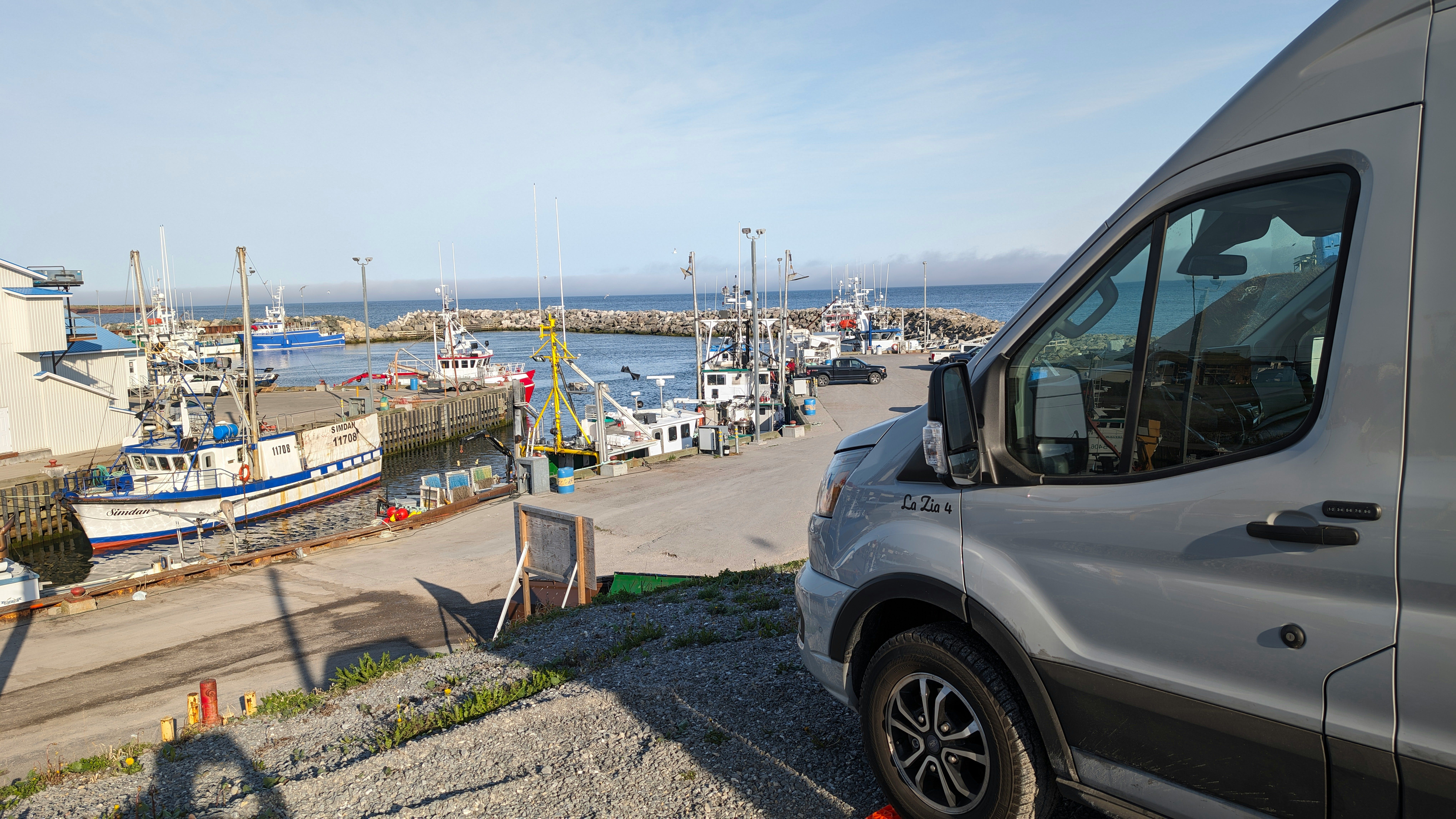 Rechts im Bild sieht man die vordere Hälfte eines grauen Kleinbusses. Er steht an einem Hafen, den man im Hintergrund sieht, mit vielen kleinen Booten. Die Sonne scheint.
