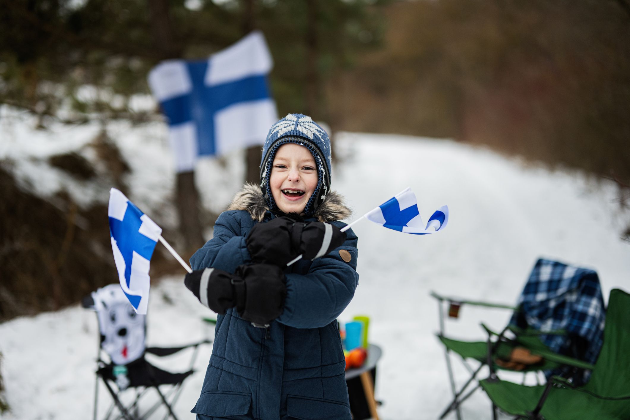 Ein Junge von ca. 6 Jahren ist warm angezogen und hat zwei Finnland-Flaggen in der Hand. Er lacht und steht in der verschneiten Natur vor ein paar Campingstühlen. Im Hintergrund sieht man eine weitere Finnland-Fahne.