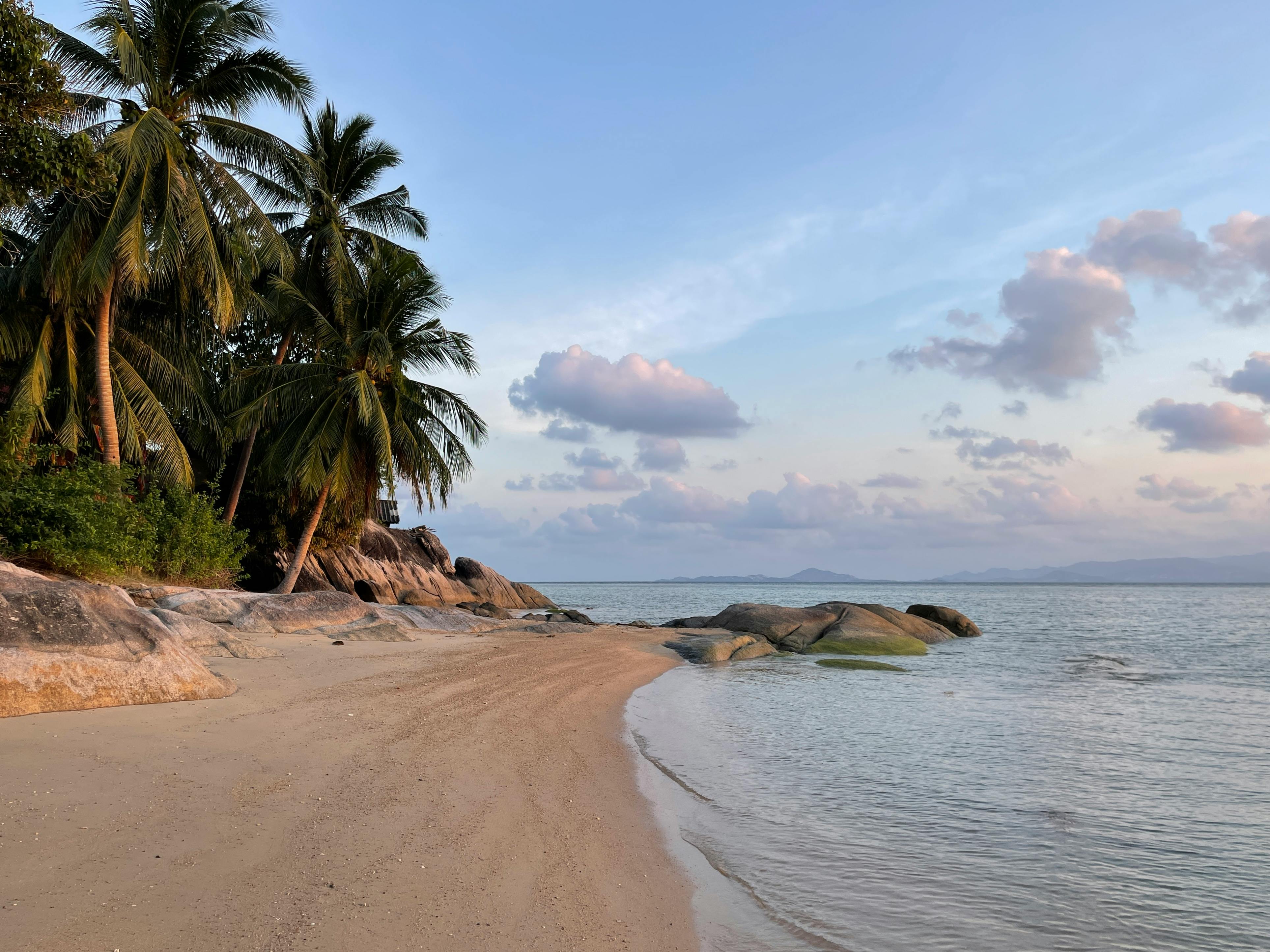 Ein einsamer Sandstrand auf Ko Samui bei Sonnenuntergang. Links im Bild sieht man Palmen, in der Mitte verläuft der Strand, und rechts liegt das ruhige Meer. Der Himmel ist leicht bewölkt.