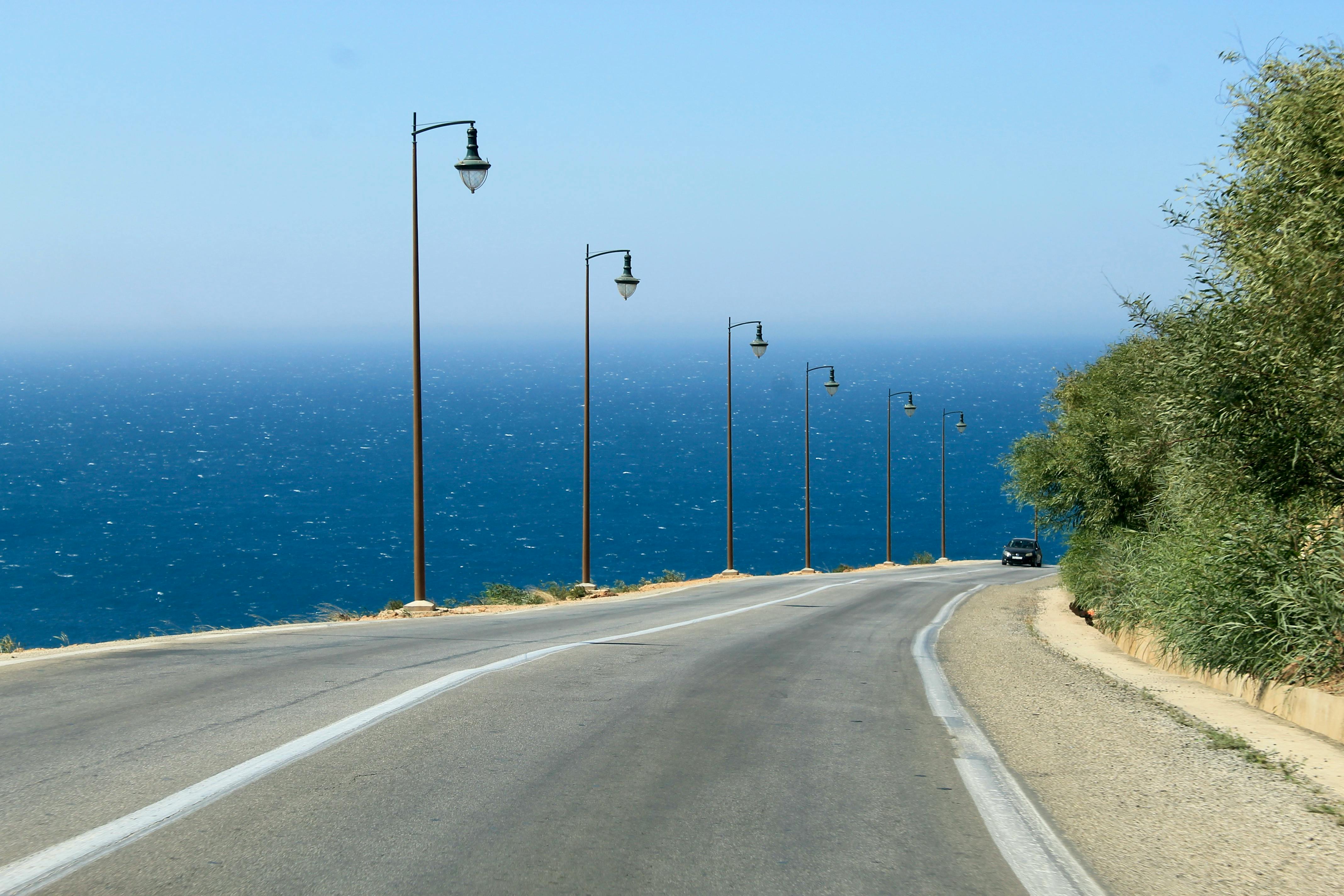 Eine Straße führt direkt an der Küste entlang. Im Hintergrund funkelt das blaue Meer in der Sonne, am Straßenrand sieht man sechs Laternen. In der Ferne erkennt man ein schwarzes Auto.