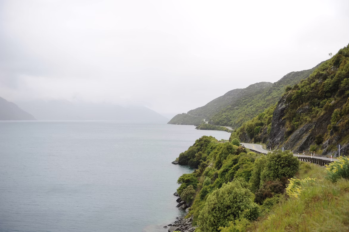 Den Großteil des Bildes nimmt der See Lake Wakatipu ein, der mystisch von Nebel umgeben ist. Rechts im Bild sieht man die sich windende Straße der Kingston Road in der Nähe von Queenstown, Neuseeland.