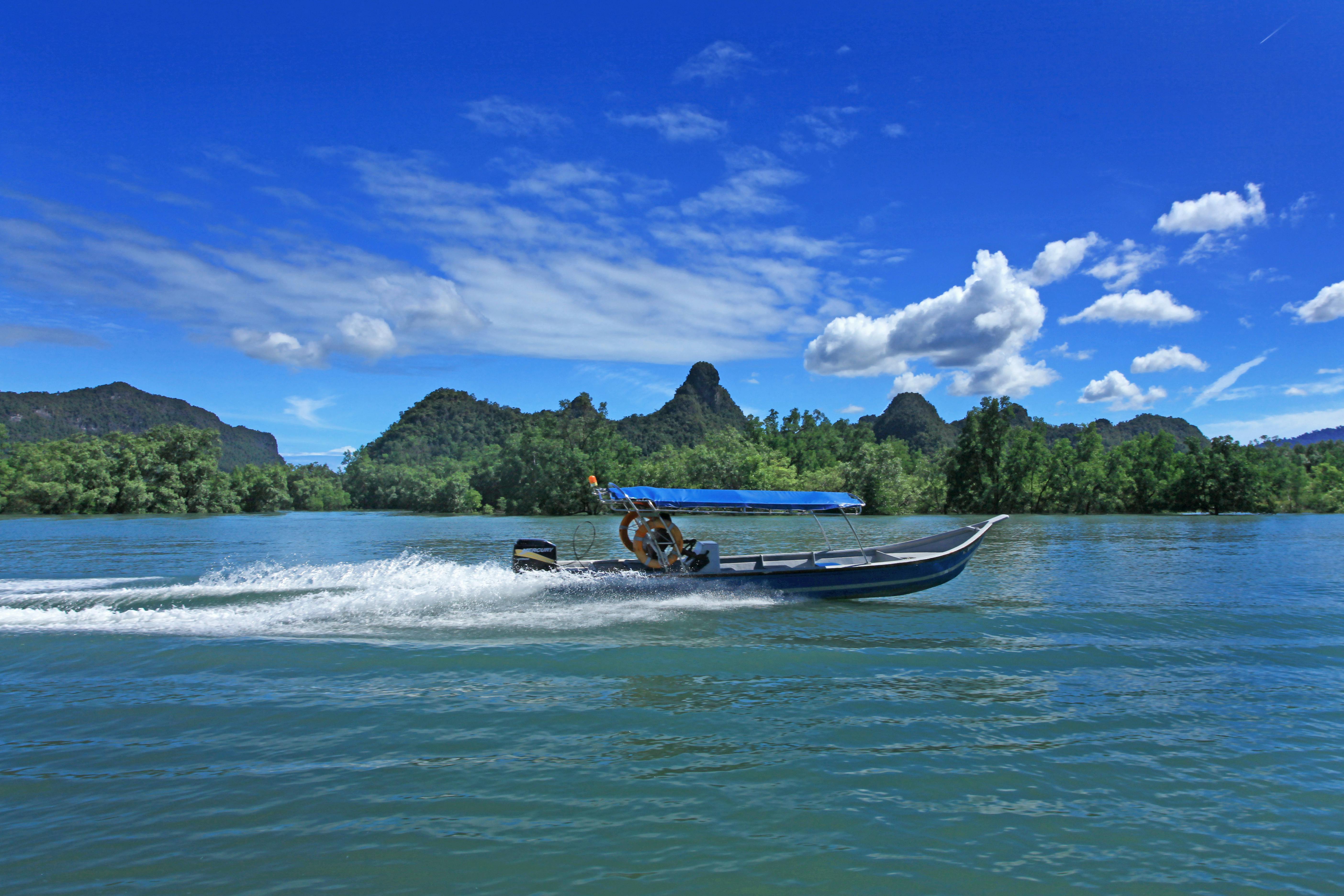Über das blaue Meer in Thailand saust ein Langboot. Im Hintergrund sieht man üppig grüne Landschaft, der Himmel ist blau und leicht bewölkt.