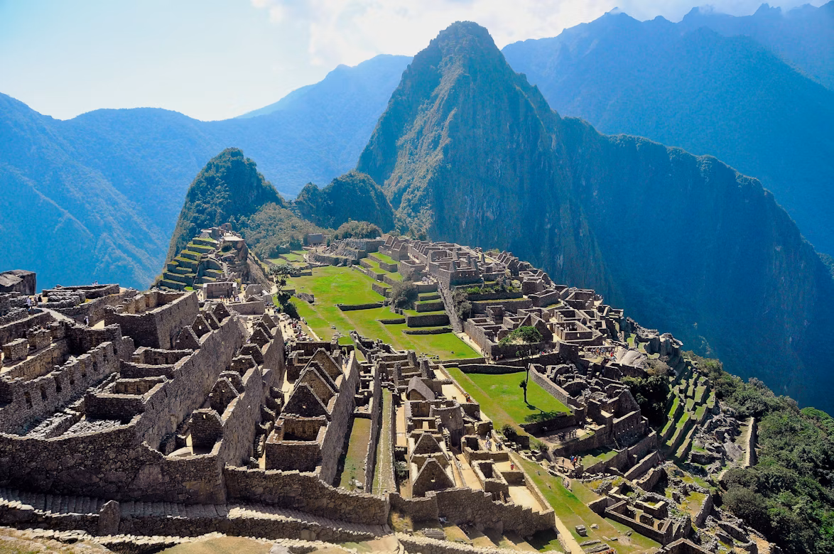 Das Foto zeigt Machu Picchu in der Sonne. Die Ruinen und das grüne Gras sind im Vordergrund, im Hintergrund sieht man beeindruckende Felsen in blauem Licht.