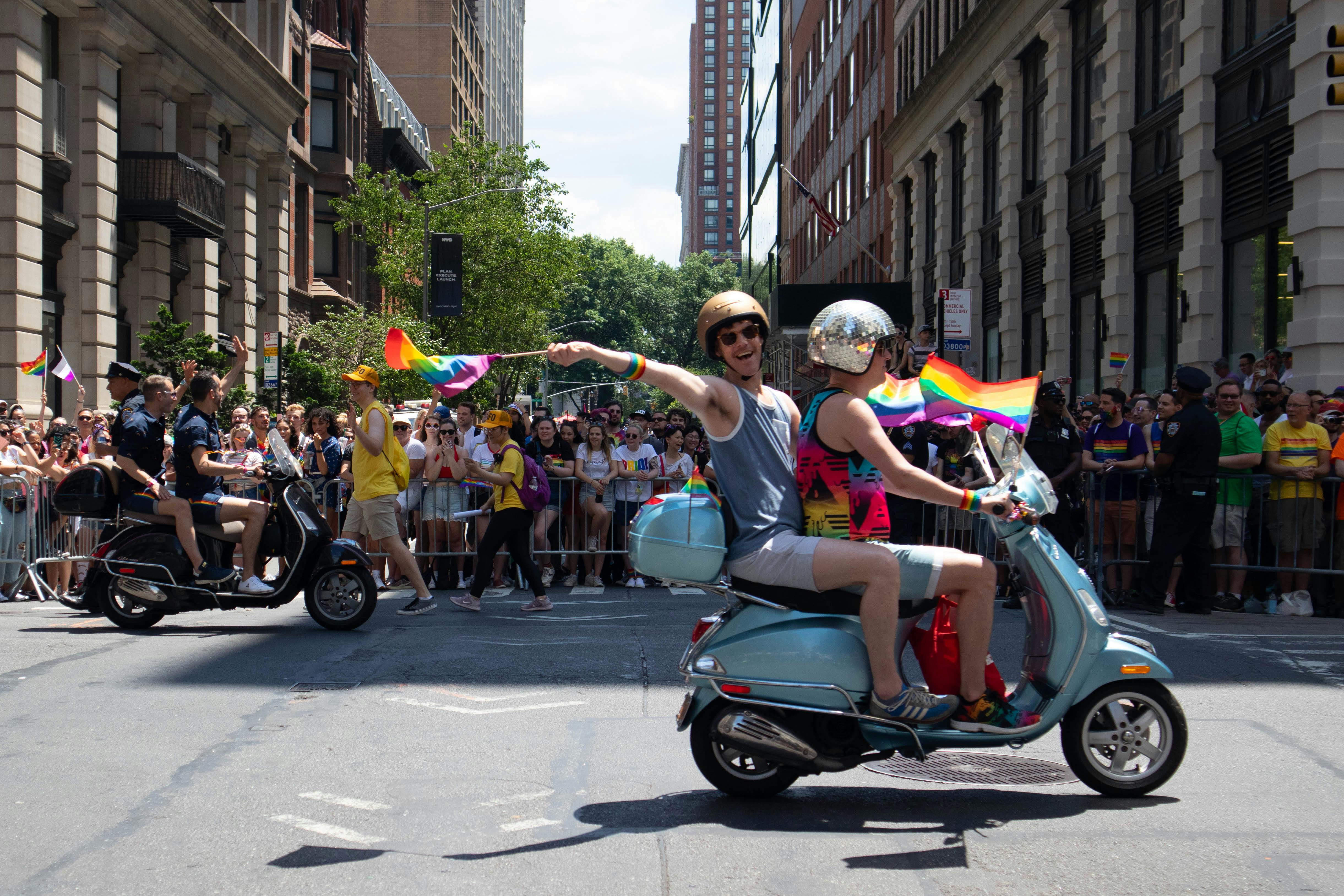 In New York fahren Menschen im Rahmen der Pride-Parade mit wehenden Regenbogenflaggen auf Scootern auf den Straßen. Im Hintergrund stehen zahlreiche Zuschauer, die die Parade anschauen.