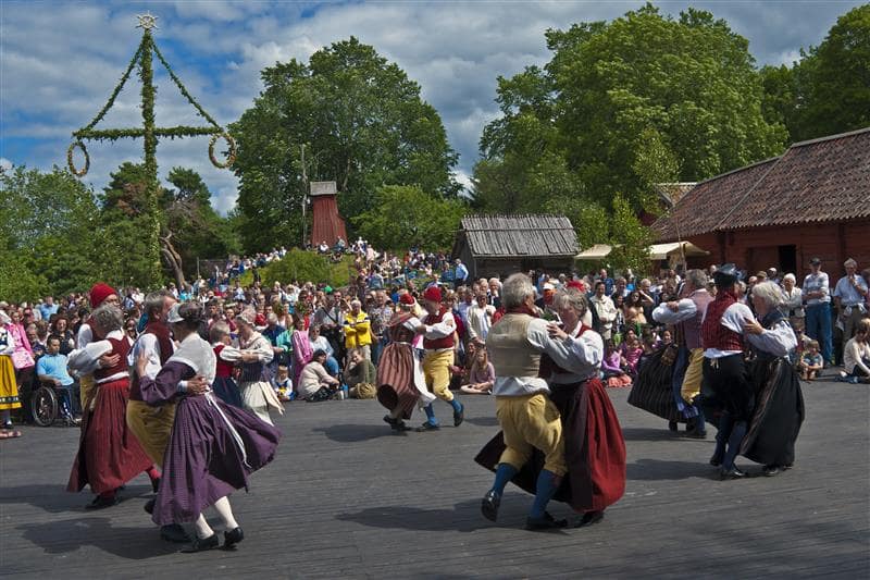 Tanzende Menschen in traditionellen Outfits. Im Hintergrund ein Midsommarbaum und Zuschauer.