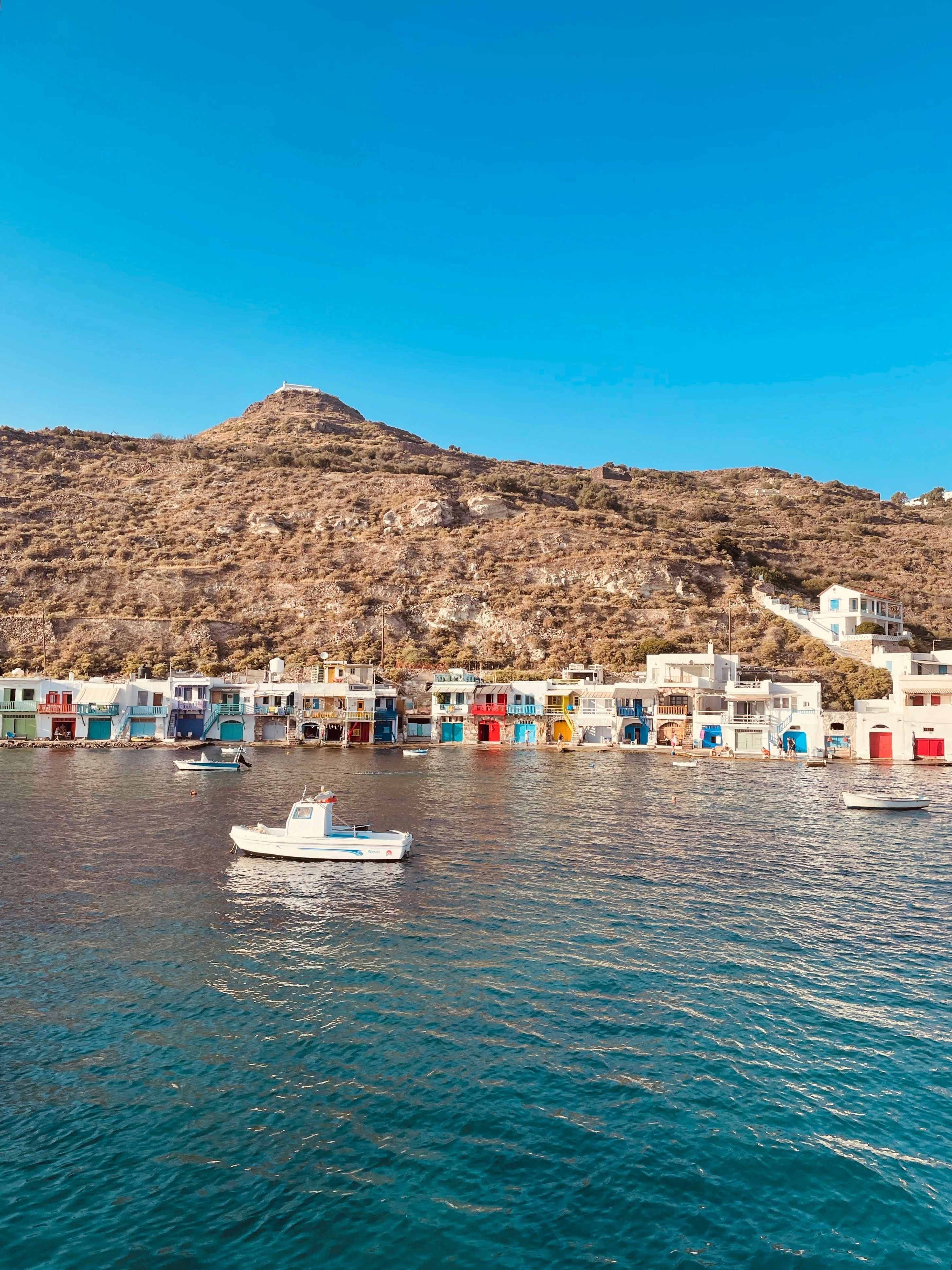 Zwischen dem blauen Himmel und blauen Meer liegt ein trockener Hügel. Am Fuße des Hügels stechen weiße Häuser mit bunten Farbakzenten heraus. Auf dem Meer sind ein paar kleine Boote zu sehen.