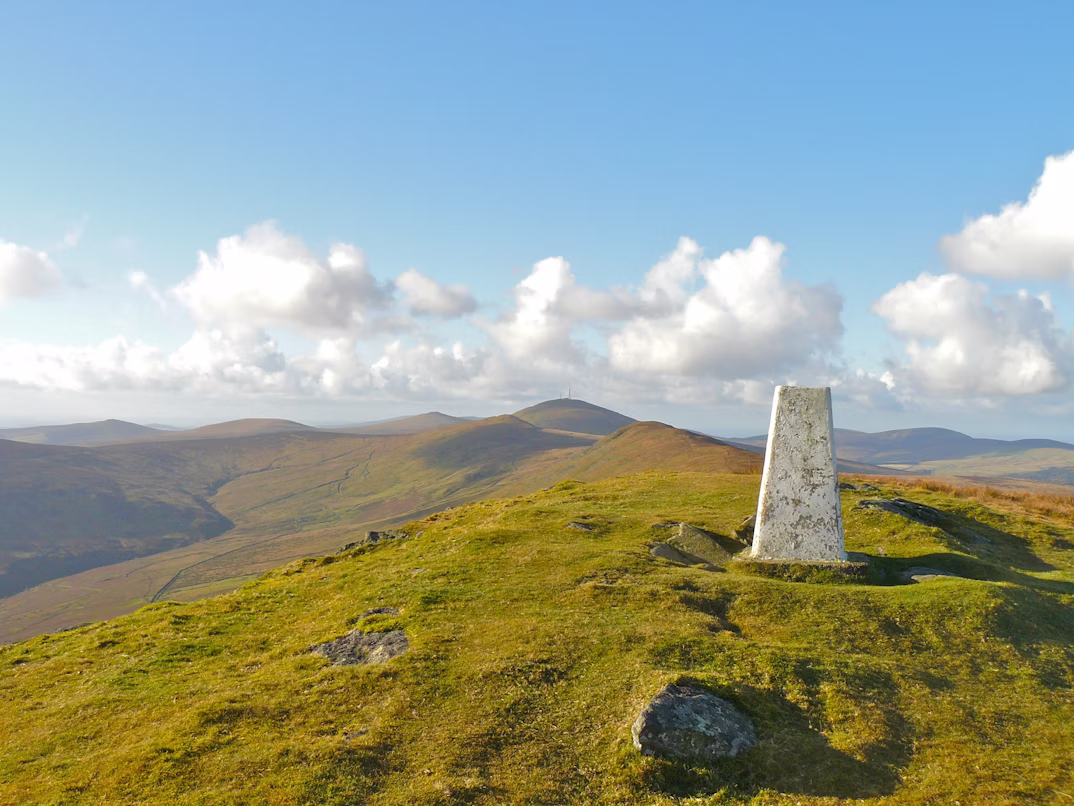 Blick über den zweithöchsten Berg der Isle of Man, den North Barrule. Das Gras ist hellgrün-gelblich, die Sonne scheint und die Sicht zeigt viele Hügel.