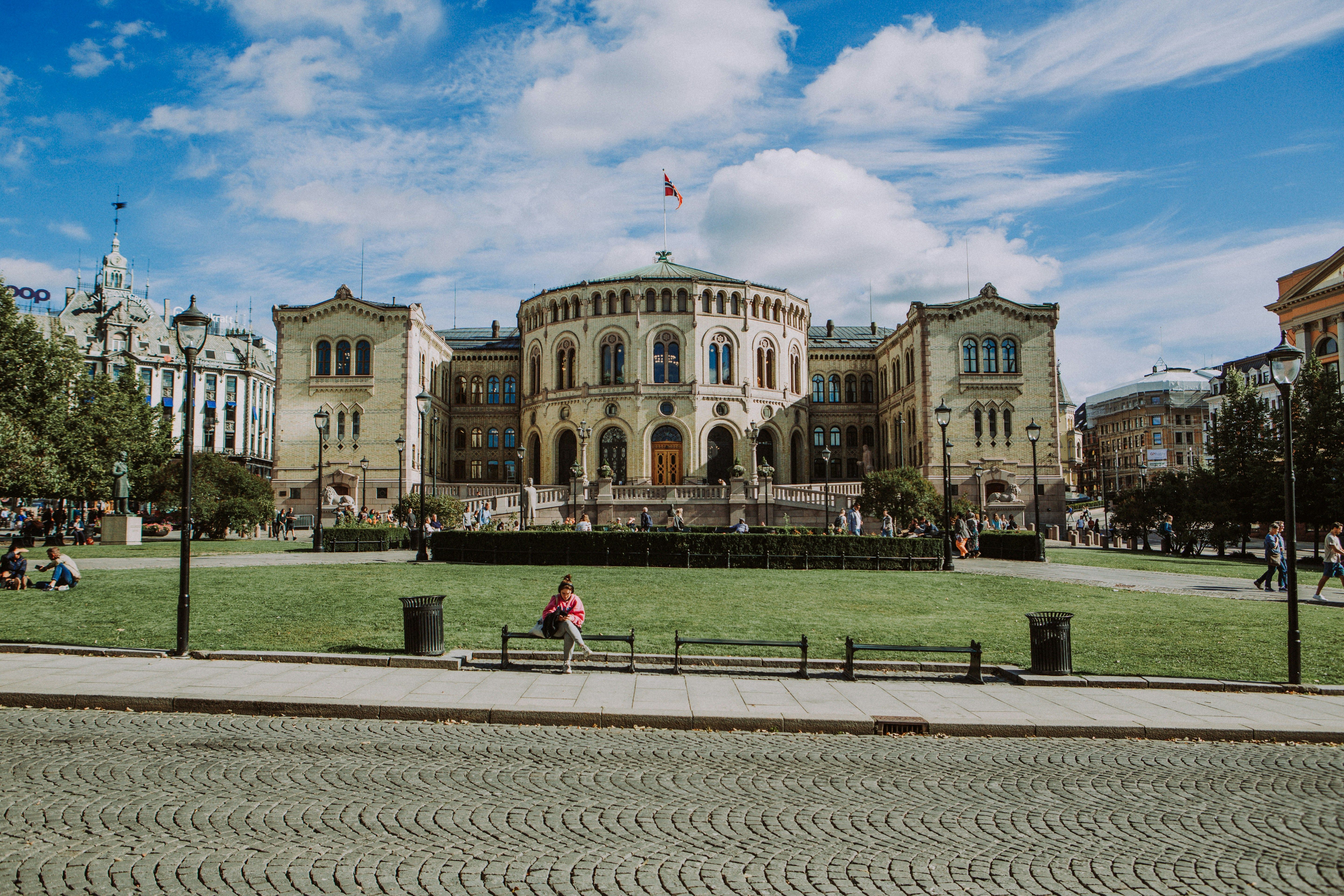 Das Parlamentsgebäude in Oslo in der Sonne. Auf dem Dach des runden Gebäudes ist die norwegische Flagge gehisst. Im Vordergrund ein akkurates Stück Rasen mit drei Bänken, auf einer sitzt eine Person.