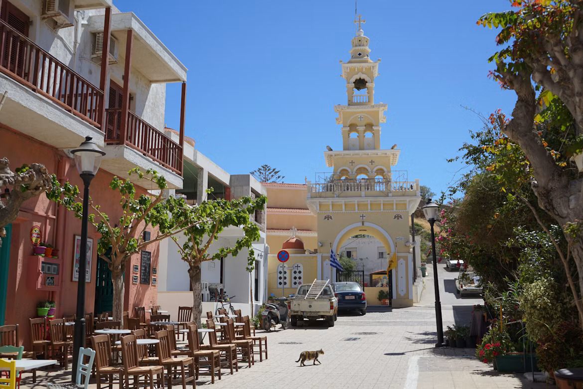 Un gato cruzando una calle soleada en Creta, con un campanario al fondo.