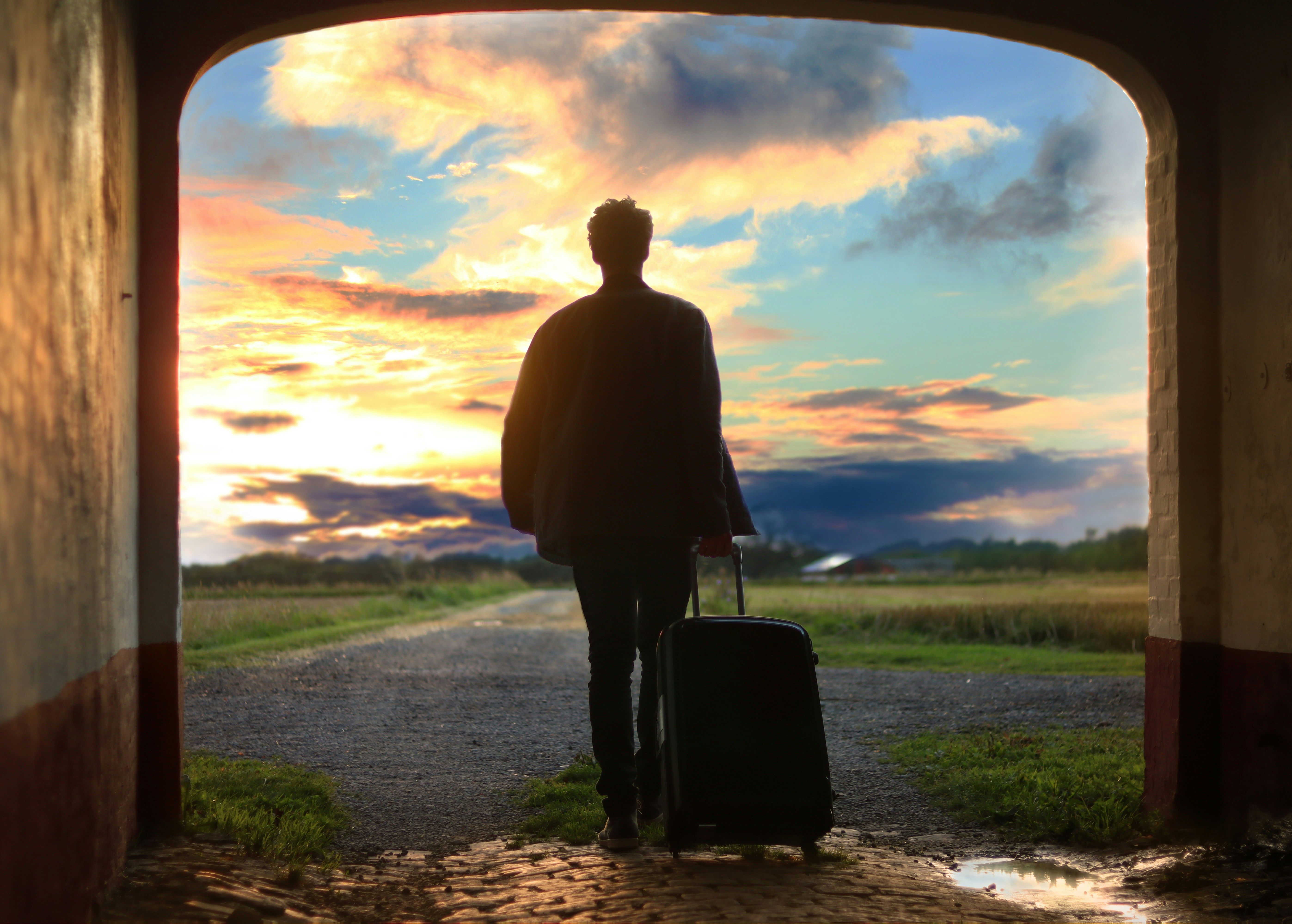 Eine Person geht bei Abendlicht über einen Weg. Die Person zieht einen Reisekoffer hinter sich her und ist nur als Silhoutte zu erkennen.