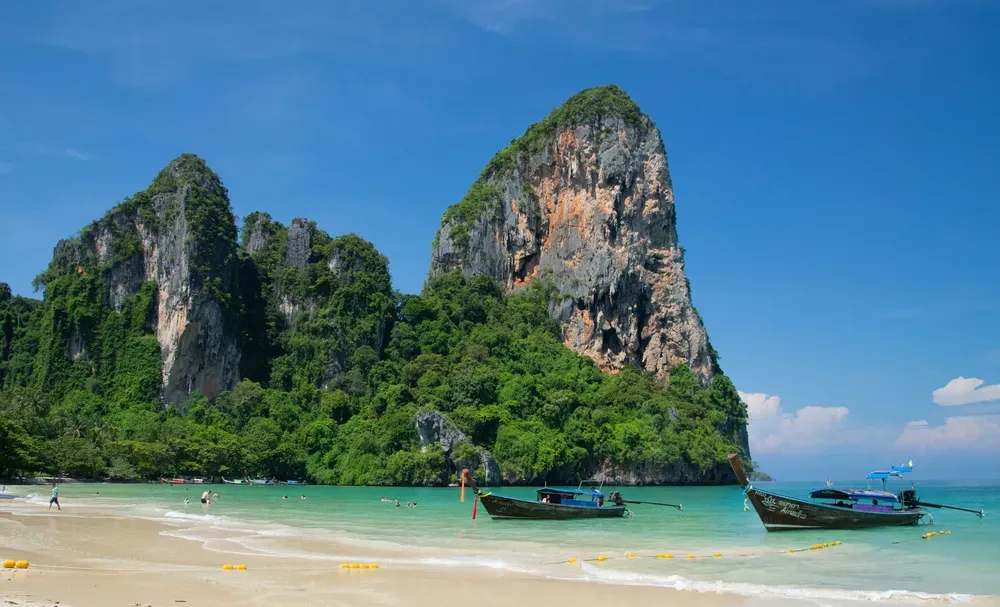 Die zerklüfteten und begrünten Felsen des Railay Beach auf Krabi ragen hinter dem weißen Sandstrand und türkisfarbenen Wasser empor.