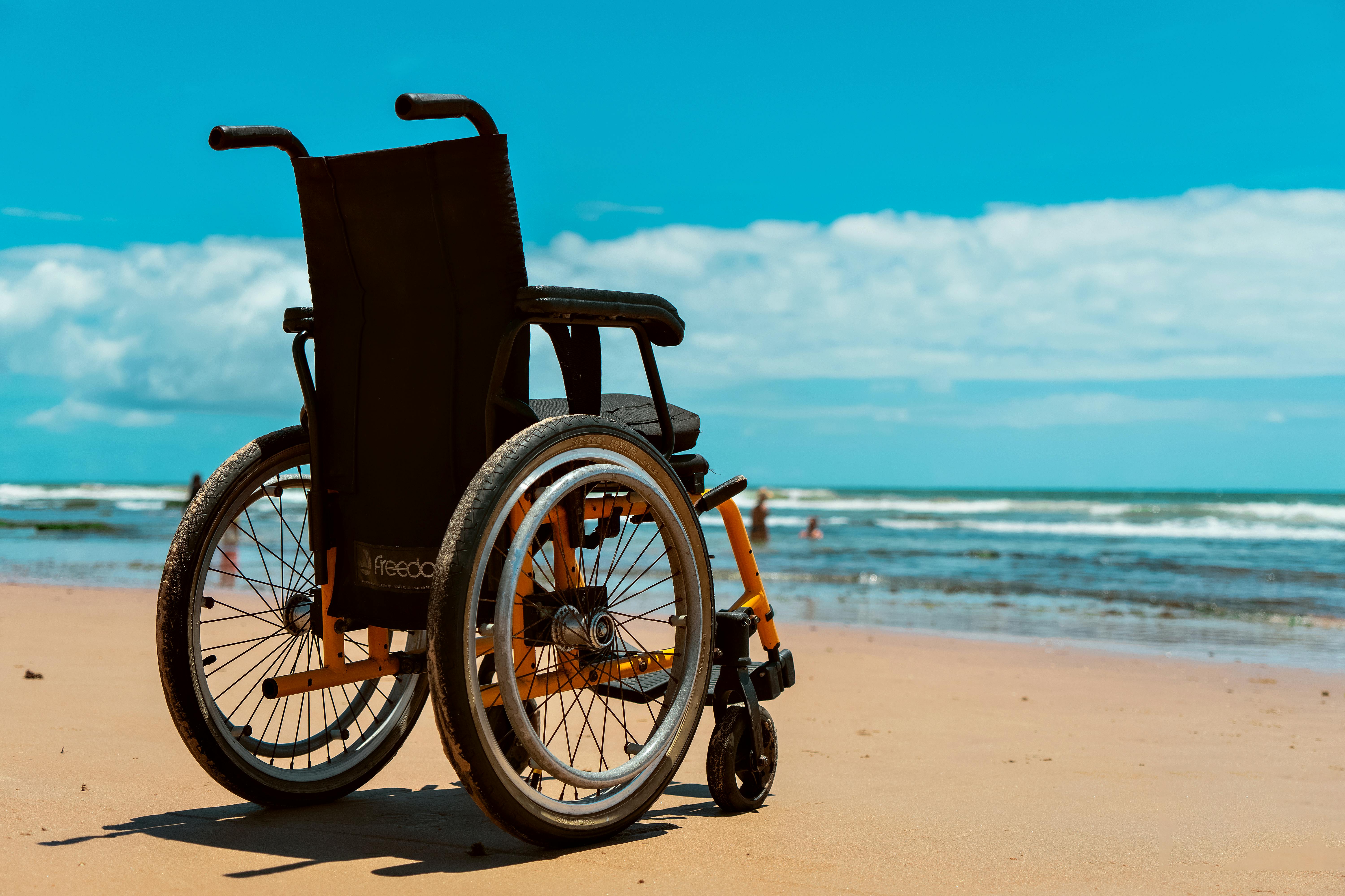 An einem Sandstrand steht ein leerer Rollstuhl. Im Hintergrund sieht man das Meer und den blauen, leicht bewölkten Himmel.