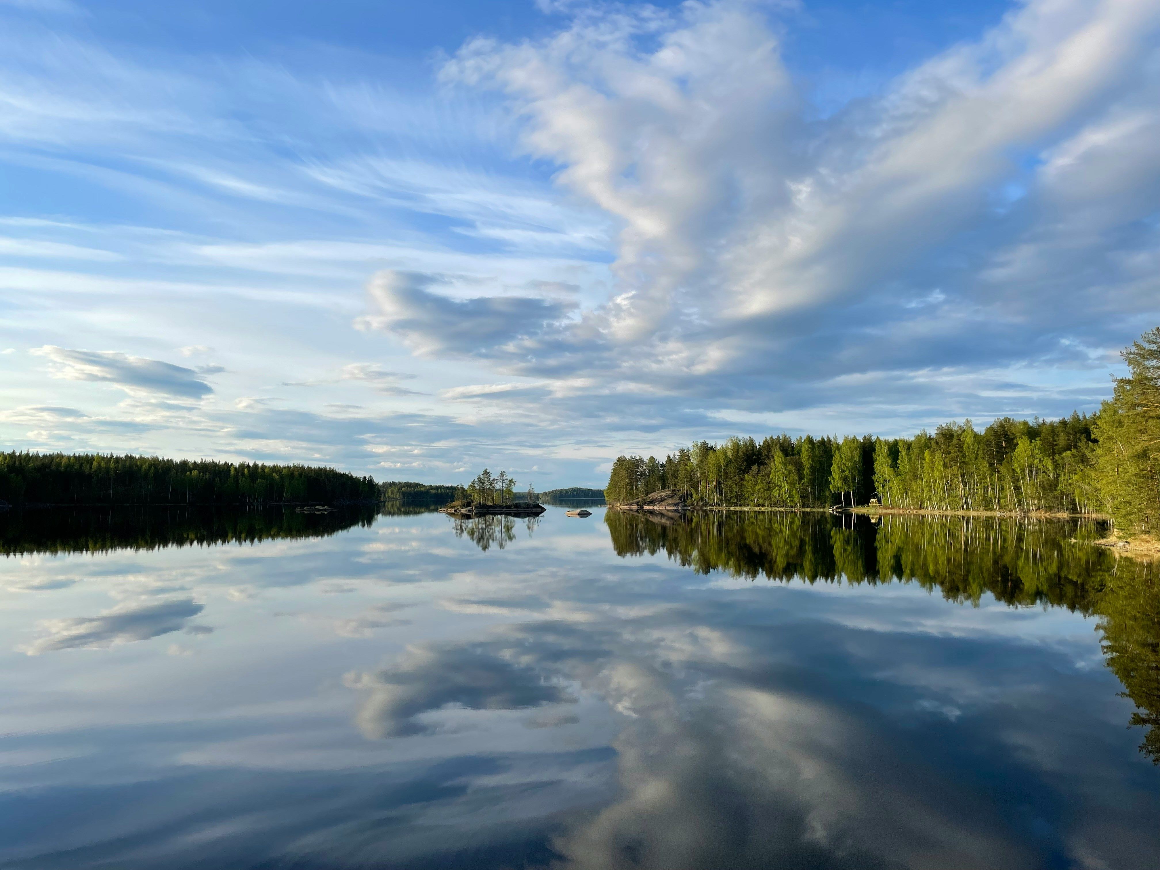 Der Saimaa-See in Finnland ist der größte See des Landes. Der leicht bewölkte Himmel spiegelt sich auf der ruhigen Wasseroberfläche, rund herum stehen Nadelbäume.