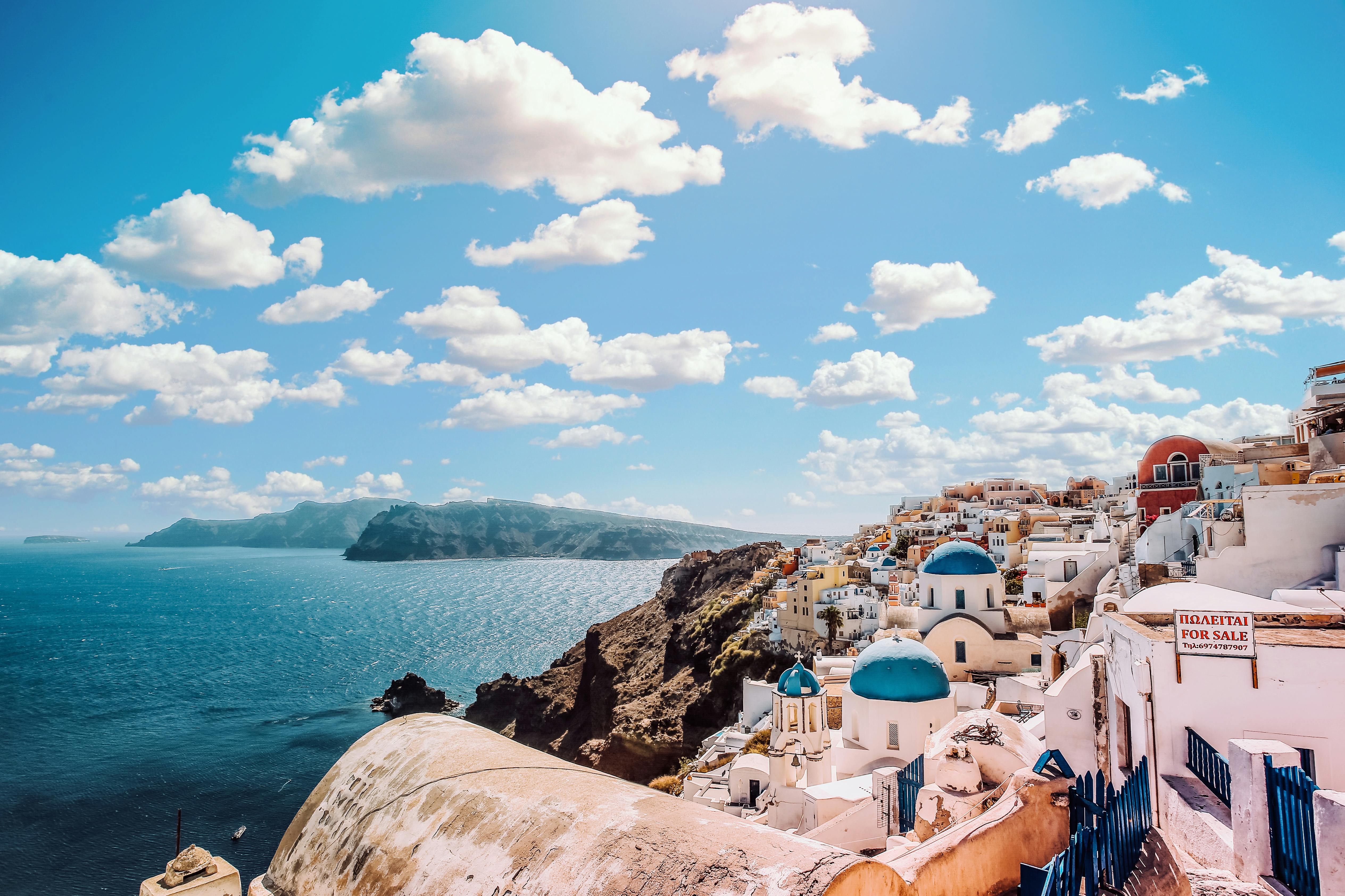 Een blik vanuit de stad op de open zee. Rechts in beeld staan de wit-blauwe huizen van Santorini. Links ligt de blauwe zee met enkele rotsen op de achtergrond. De lucht is blauw met een paar wolken.