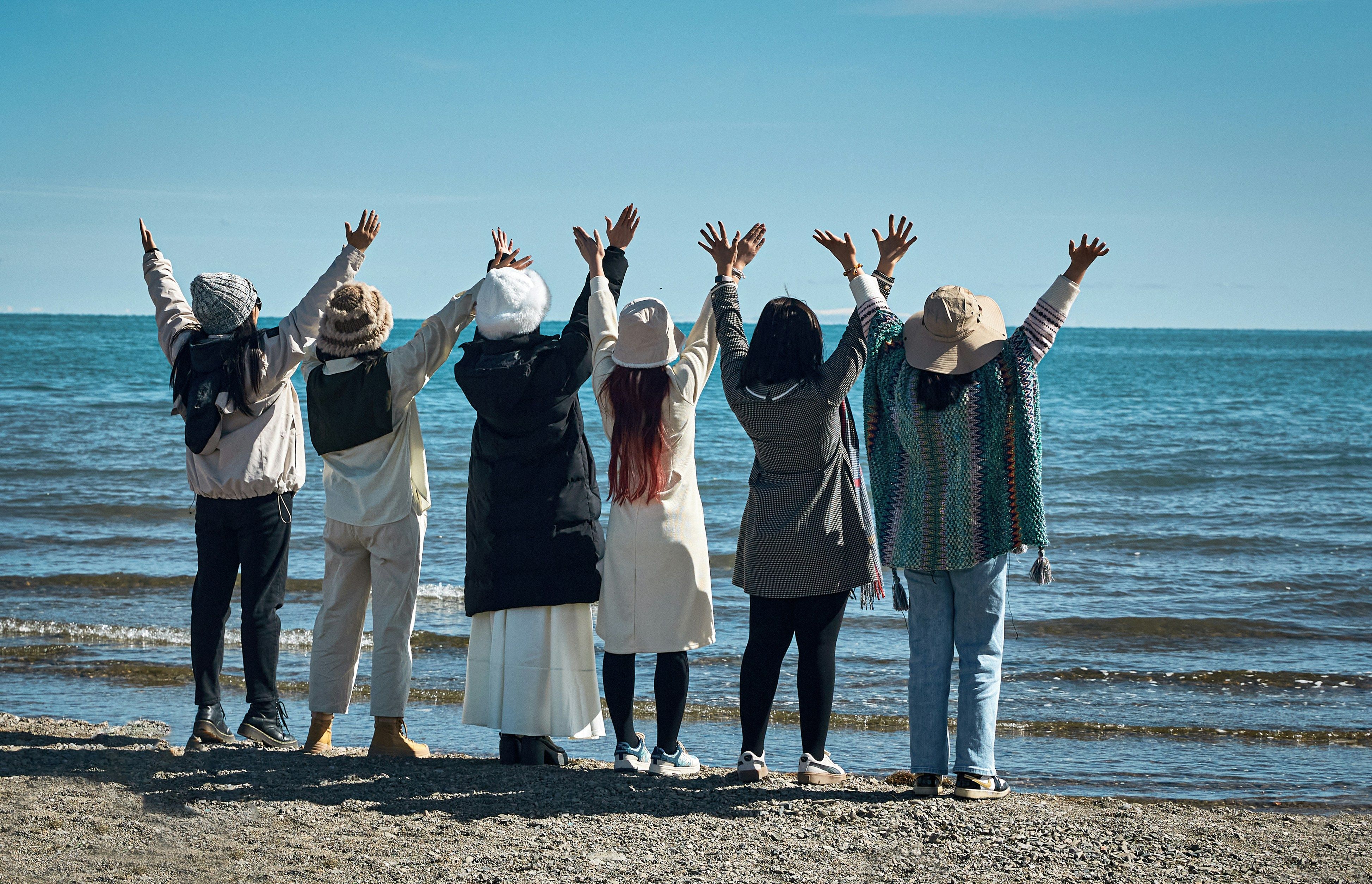 Sechs Menschen sind von hinten fotografiert. Sie stehen am Ufer des Meeres und heben alle ihre Arme in die Höhe.