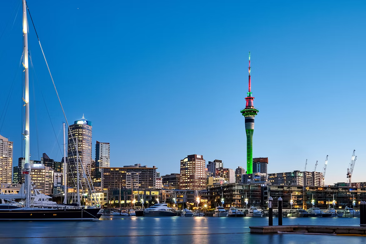 Blick auf die Skyline von Auckland, Neuseeland, vom Wasser aus. Die Sonne ist fas untergegangen, und der Sky Tower ist grün beleuchtet.