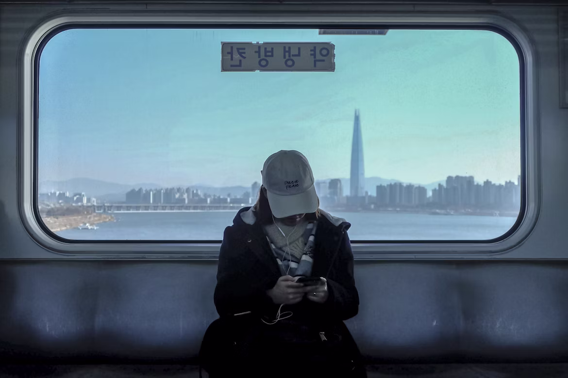 A passenger wearing a hat commuting on a train with the city of Gangnam in the distance