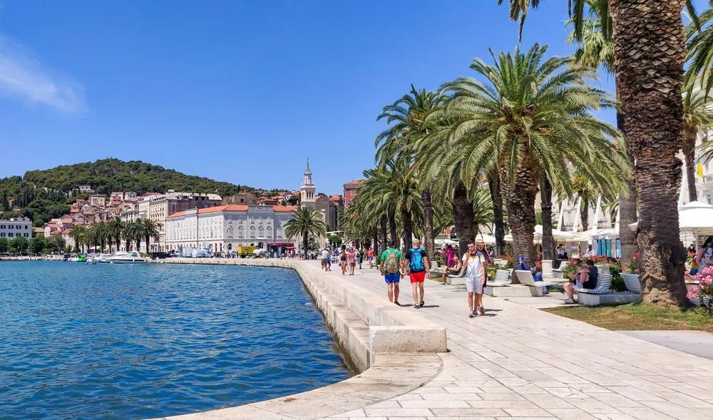 Promenade von Split. Blaues Wasser, Palmen links und laufende Menschen, im Hintergrund grüner Hügel und Gebäude.
