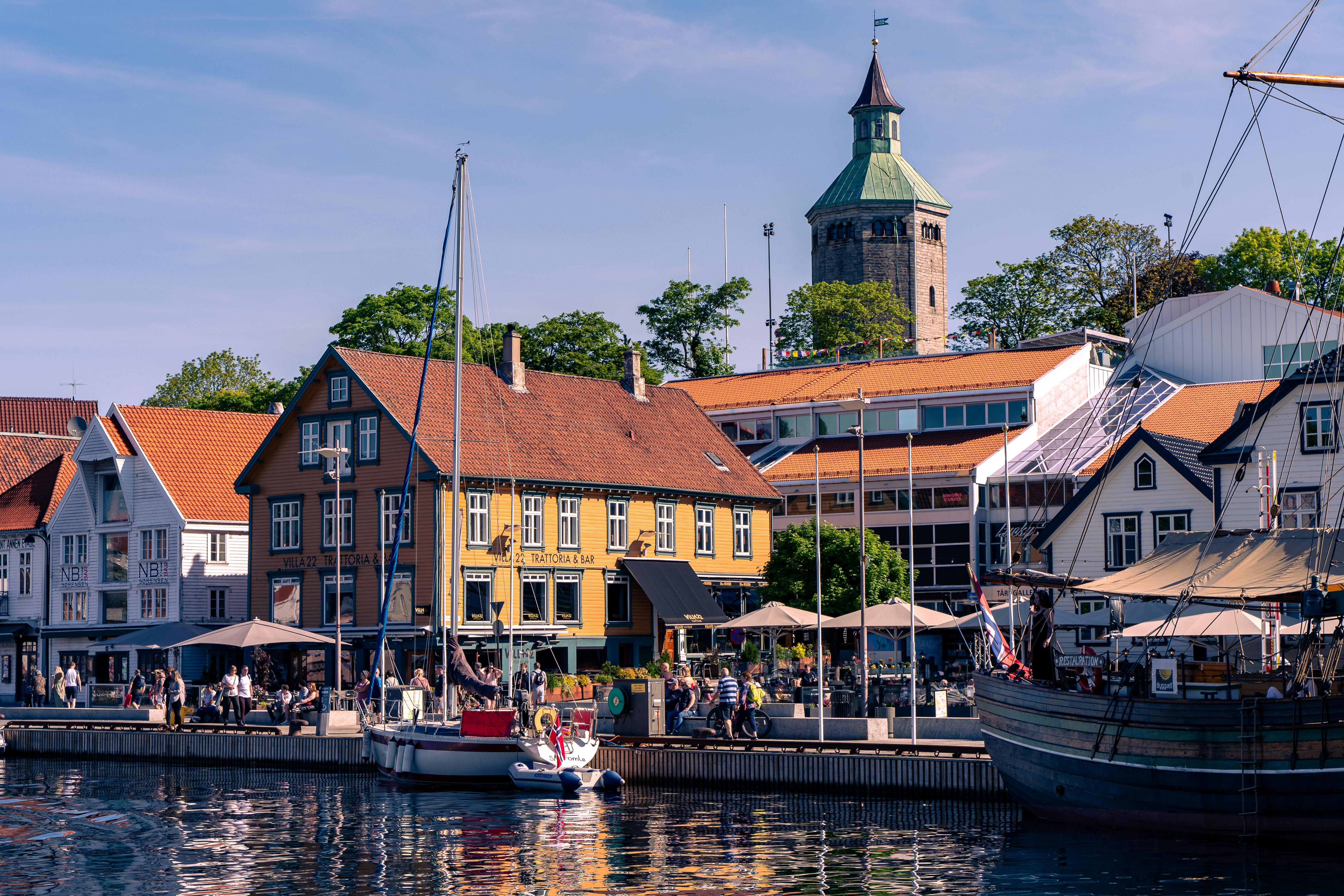 Dans la vieille ville de Stavanger, en Norvège, des restaurants bordent une promenade qui longe le front de mer. Le soleil brille et quelques bateaux de pêche naviguent sur l'eau.