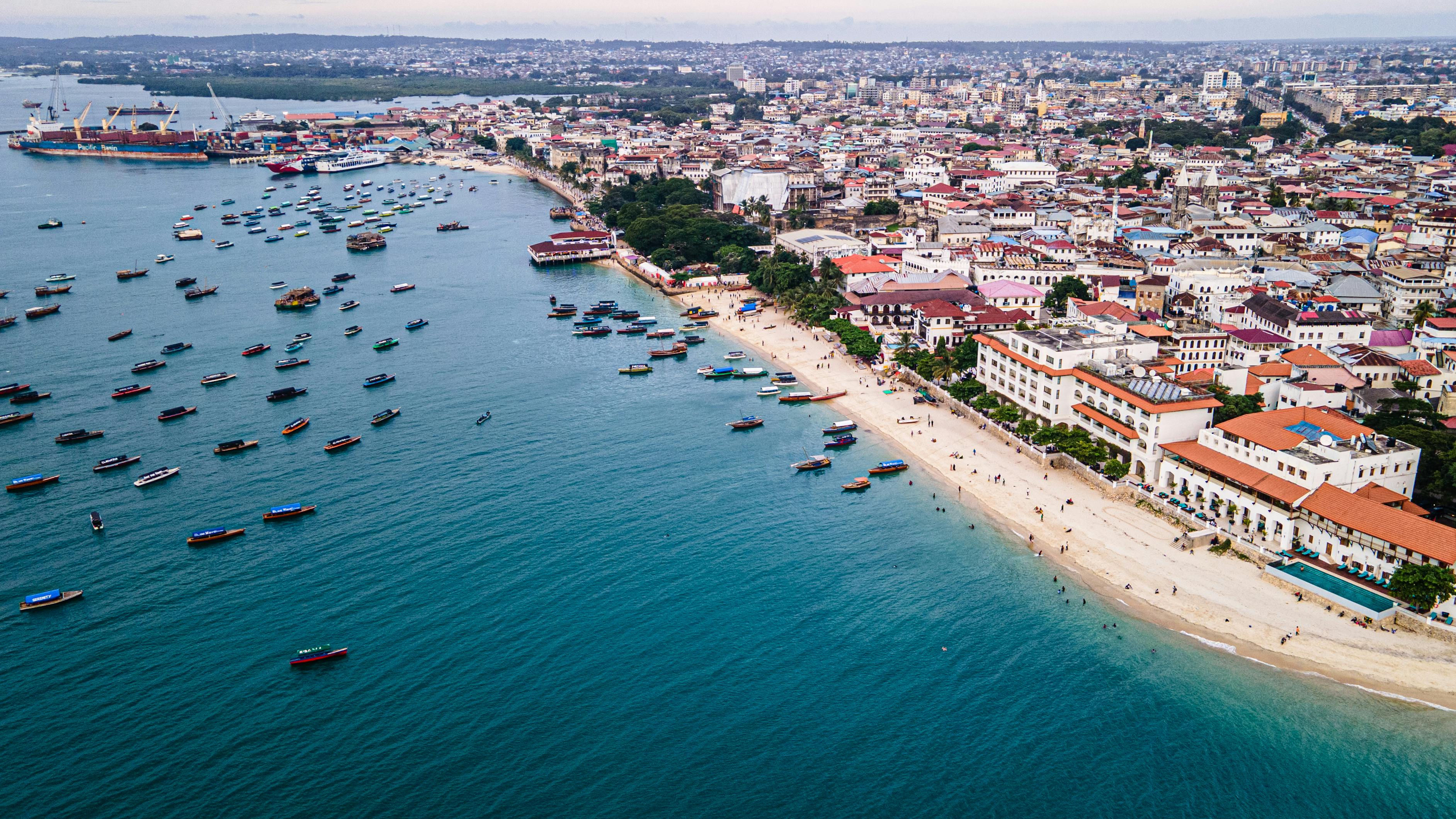 Aus der Vogelperspektive sieht man Boote im Meer, gleich vor dem Ufer eines Sandstrandes. Hinter dem Strand liegt eine Stadt mit Kolonialarchitektur. Es handelt sich um Sansibar-Stadt auf Sansibar.