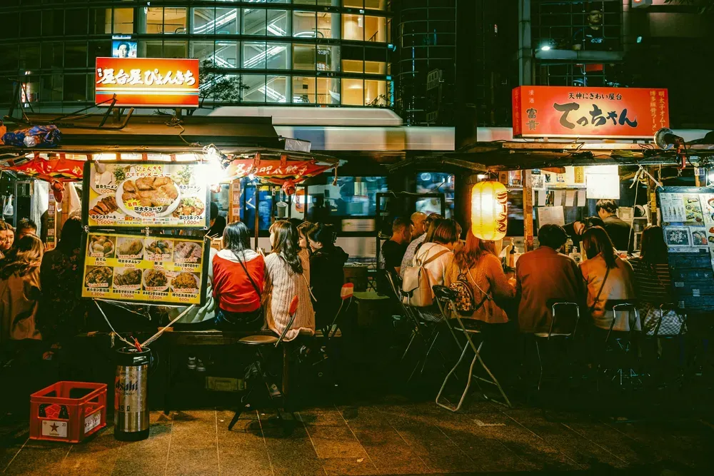 Am Abend sitzen einige Menschen zusammen an beleuchteten Streetfood-Ständen.