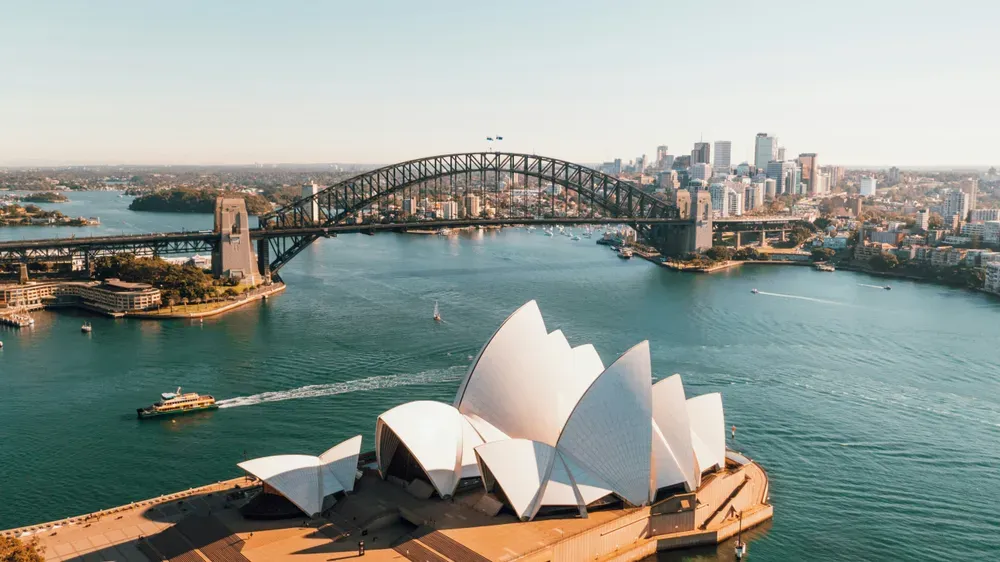 Aus der Luft fotografiert, sieht man das Opernhaus in Sydney und die dahinter liegende Harbour Bridge. Auf dem ruhigen Wasser fährt eine Fähre, die Sonne scheint.