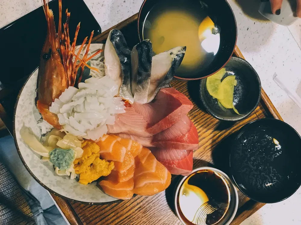 Roher Fisch und Meeresfrüchte, Reis und Beilagen liegen in einer Schüssel, die auf einem Tablett steht. Des weiteren stehen auf dem Tablett einige leere Schüsseln und eine Suppe.