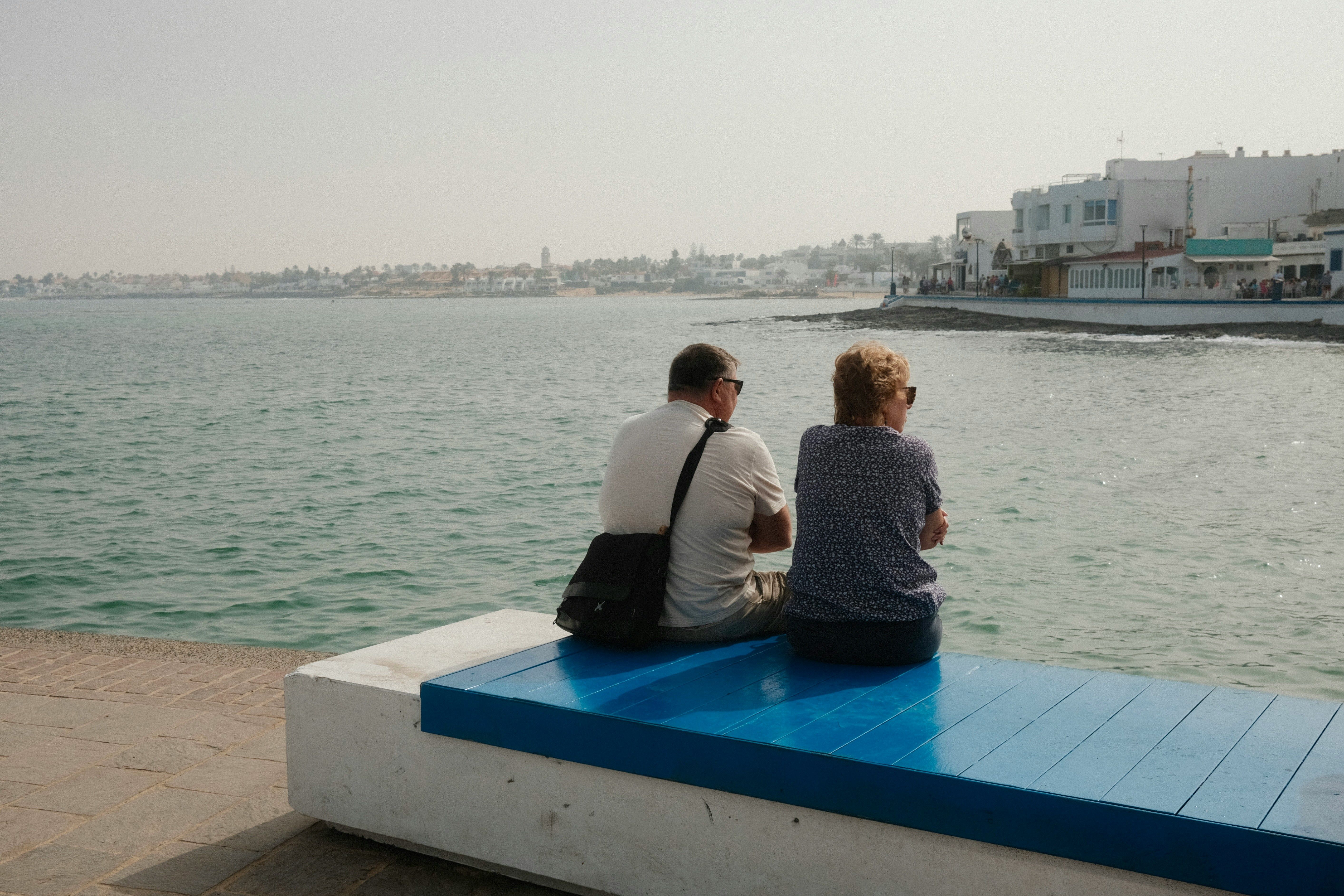 Un homme et une femme sont assis sur un banc bleu et blanc le long d'une promenade en bord de mer et regardent l'eau.