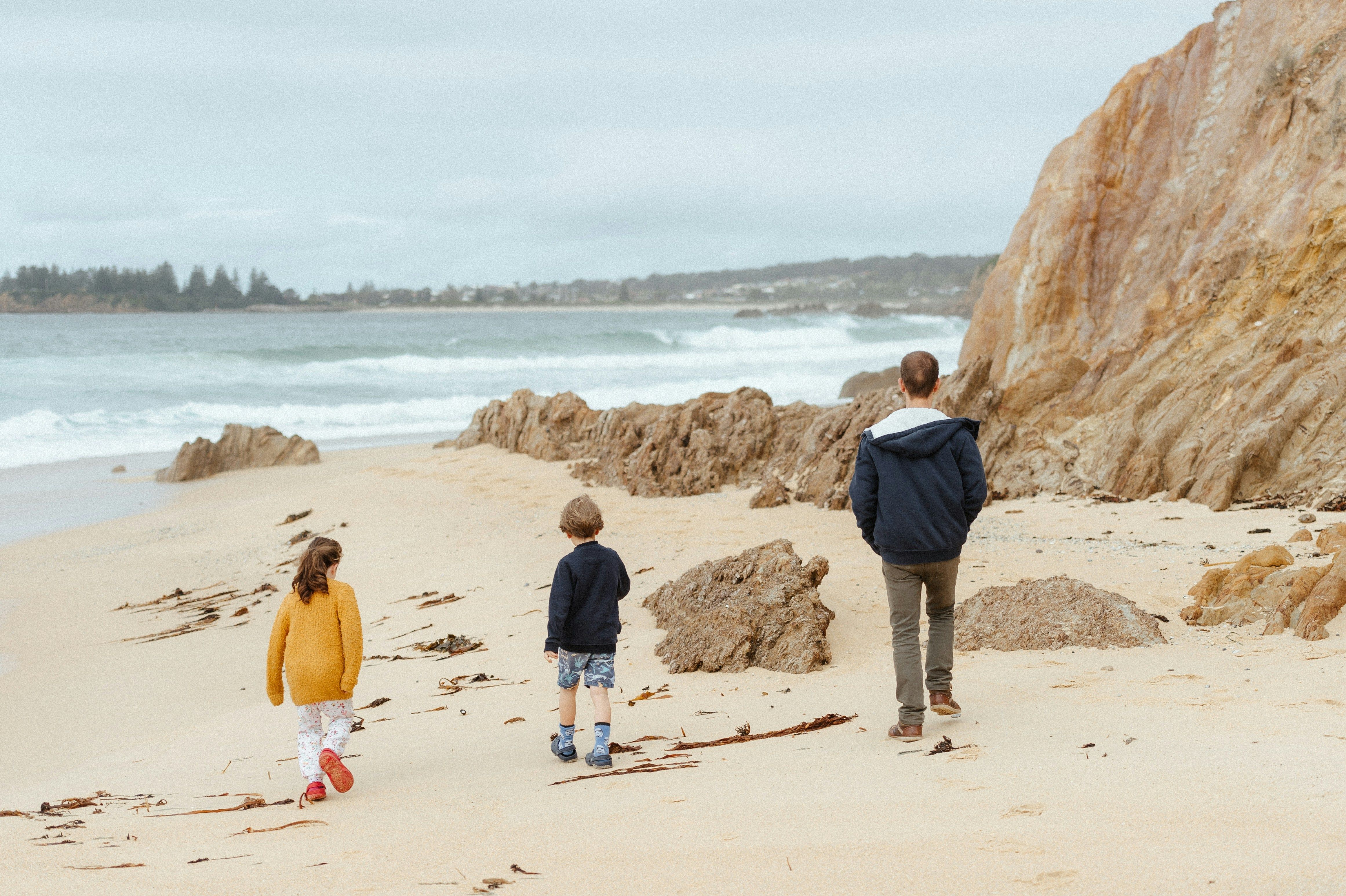 Zwei Kinderm, circa 6 Jahre alt, und ein Mann gehen am Strand spazieren. Es ist Nebensaison, alle sind etwas wärmer angezogen und das Meer im Hintergrund sieht unruhig aus.