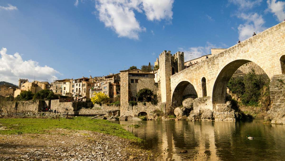 Ponte medievale di Besalú riflesso sul fiume Fluvià, con le case del centro storico sullo sfondo sotto un cielo azzurro.
