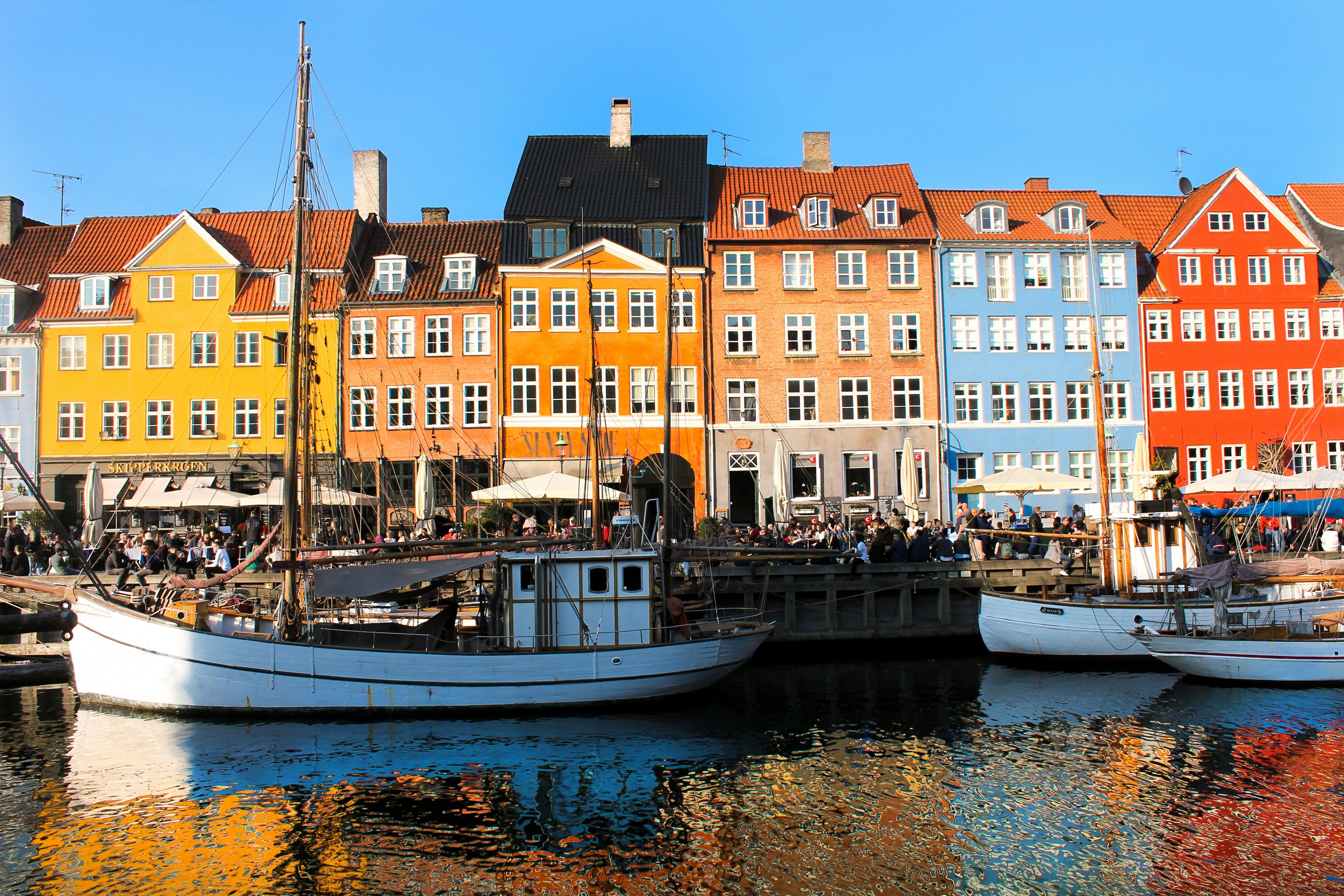 Die bunten Häuser des Nyhavn-Viertels in Kopenhagen liegen entlang eines Kanals. Der Himmel ist blau, man kann viele Menschen auf der Promenade erahnen. Auf dem Wasser sind ein paar angeleinte Fischerboote.